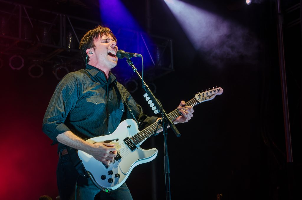Jim Adkins of Jimmy Eat World playing guitar and singing with red, blue, and purple lights surrounding him.