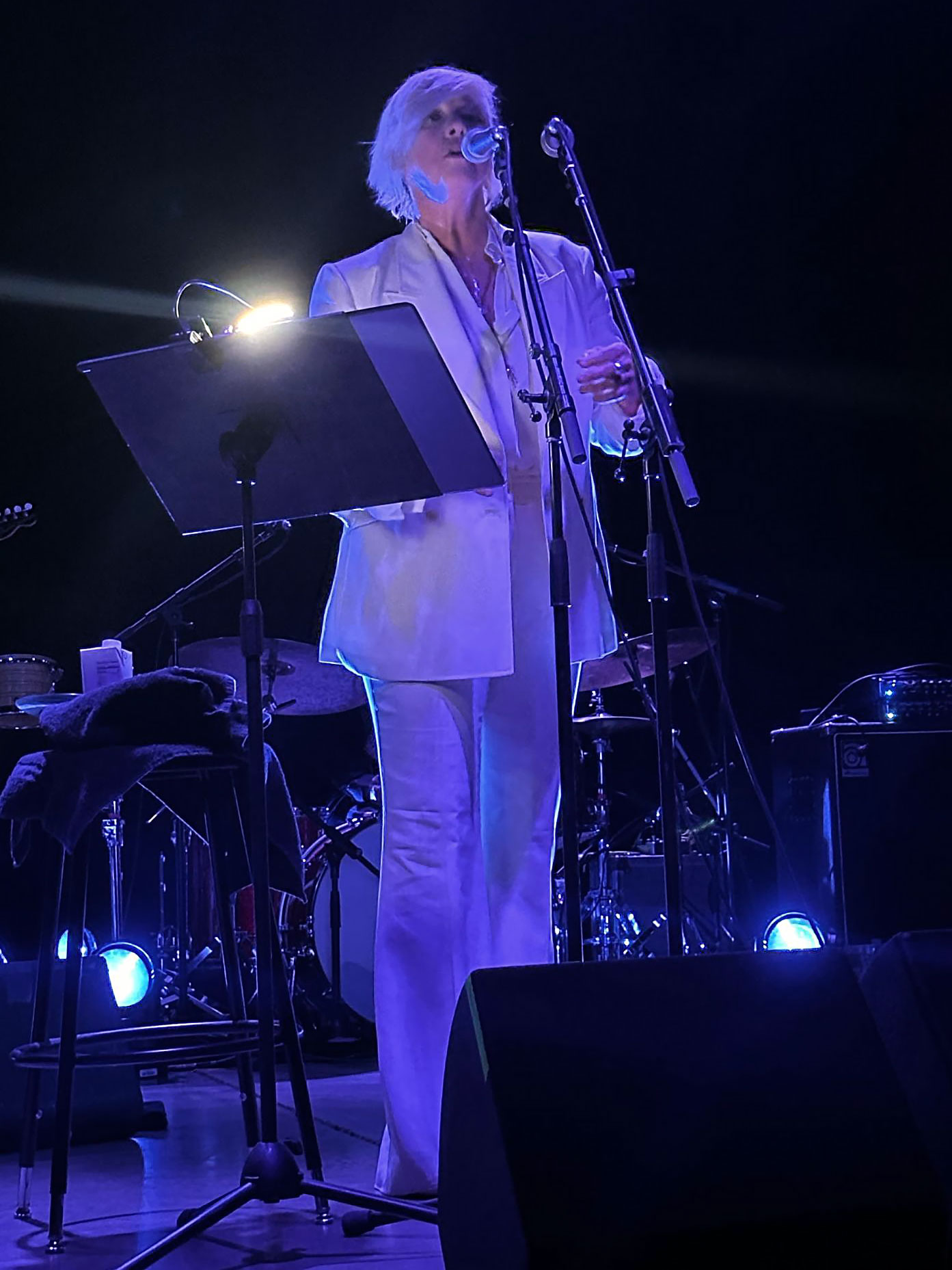 Cat Power stands at a microphone at The Van Buren in Phoenix, wearing a bright white suit under blue stage lighting. A music stand and microphone stand are positioned in front of her, with drums visible in the background.