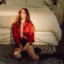 LØLØ sits on a leopard-print rug in front of a cream-colored bed, wearing a red jacket, black shorts and a silver statement top, in a promotional portrait for her “god forbid a girl spits out her feelings” era.