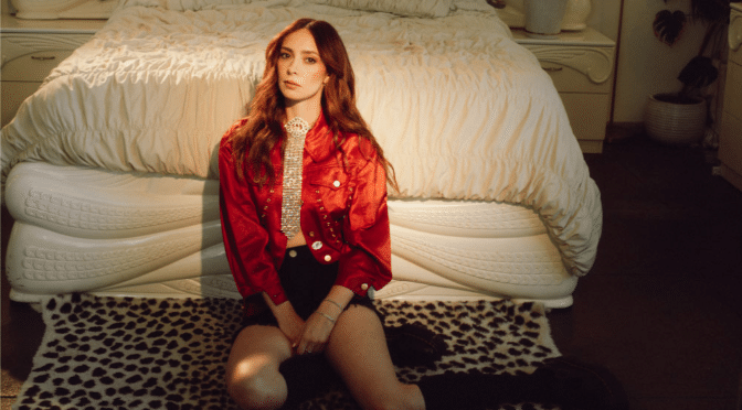 LØLØ sits on a leopard-print rug in front of a cream-colored bed, wearing a red jacket, black shorts and a silver statement top, in a promotional portrait for her “god forbid a girl spits out her feelings” era.