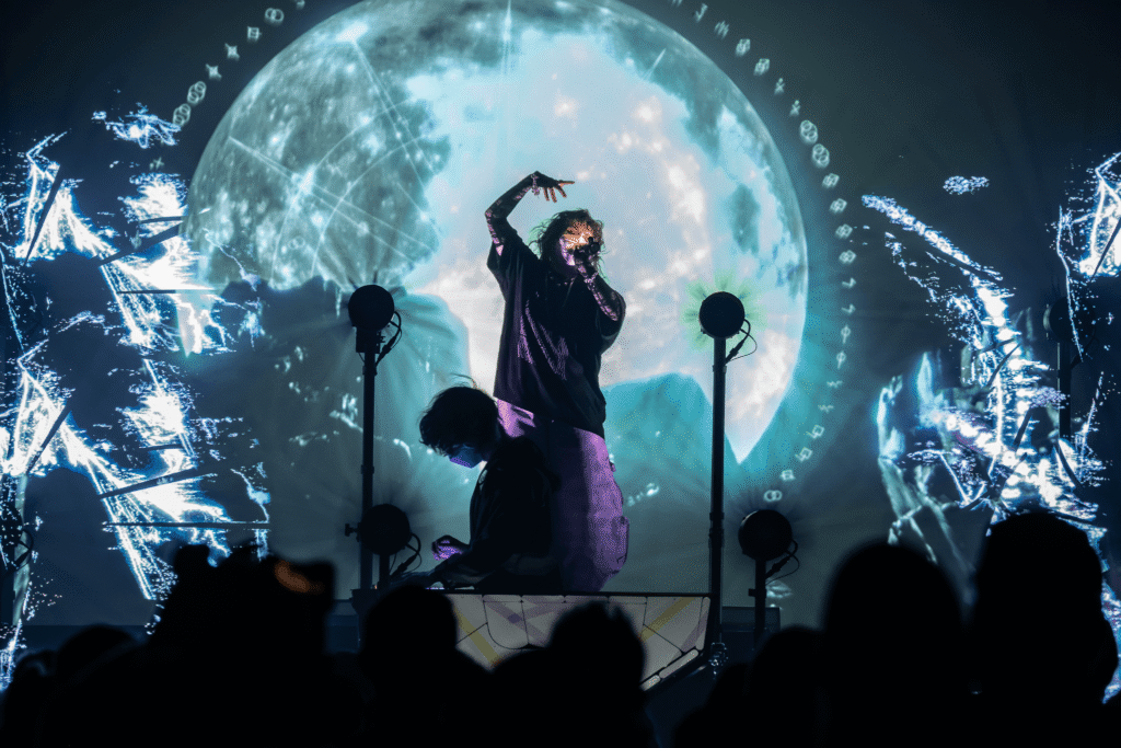 Purity Ring perform in Phoenix on their 2025 tour, against a full moon and cosmic digital visuals, captivating the crowd with immersive light and sound.