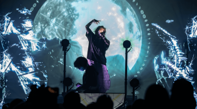Purity Ring perform in Phoenix on their 2025 tour, against a full moon and cosmic digital visuals, captivating the crowd with immersive light and sound.