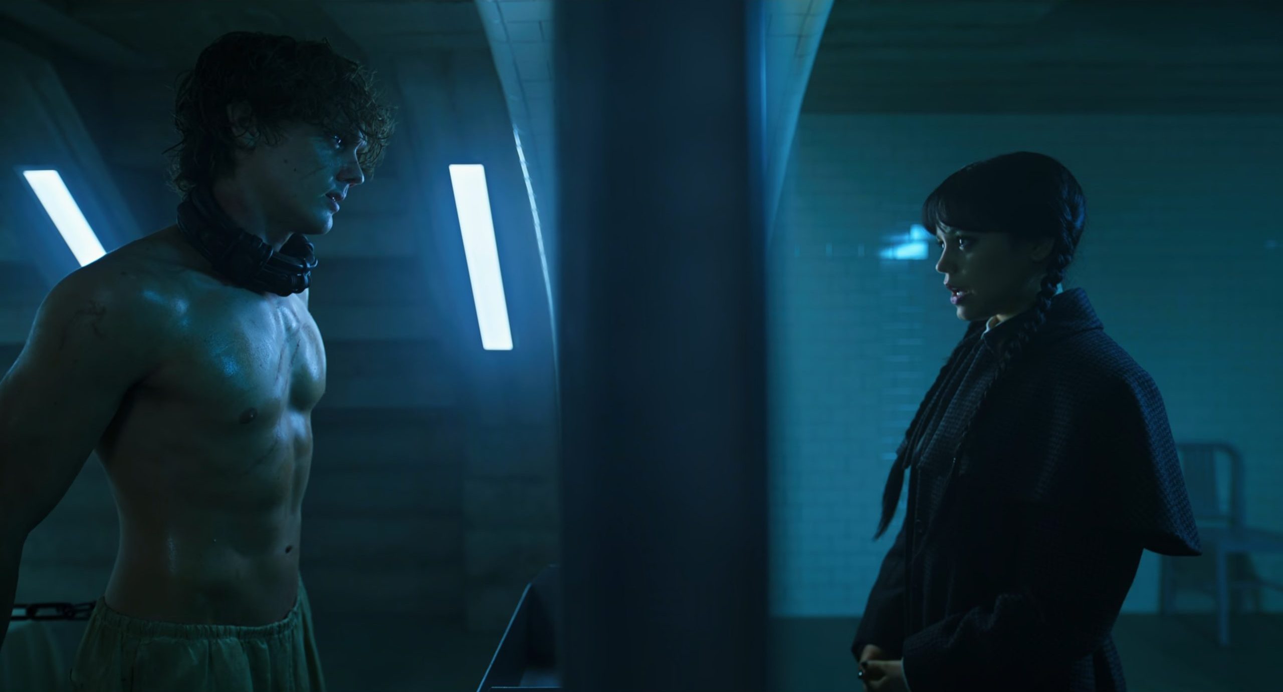 Tyler Galpin shirtless and chained, facing Wednesday Addams in a dimly lit, blue-toned underground room. The characters are separated by a pillar, highlighting the tension between them.