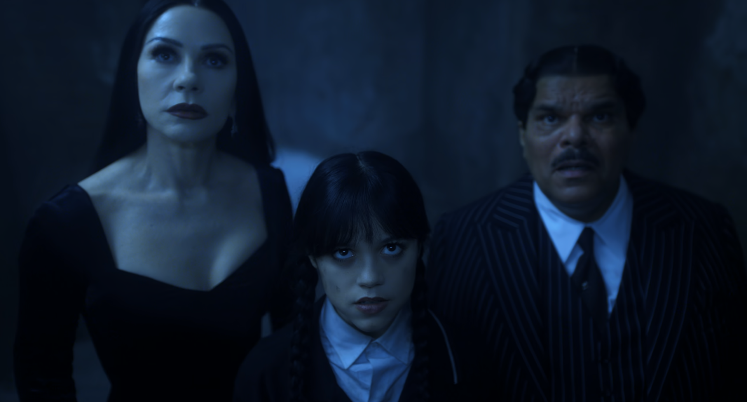 Wednesday Addams stands between Morticia and Gomez, all three looking upward with tense, serious expressions in a dim, blue-lit scene. Morticia wears a black dress, Gomez is in a pinstripe suit, and Wednesday stares intensely ahead with her signature braids.