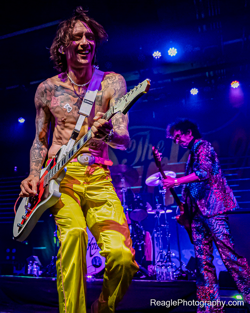 Justin Hawkins smiling widely as he plays the guitar, with Frankie Poullain playing bass in the background