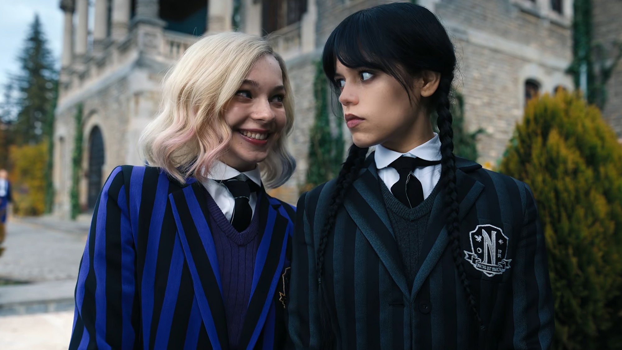 Enid Sinclair smiling wide at resting-faced Wednesday Addams outside Nevermore Academy.