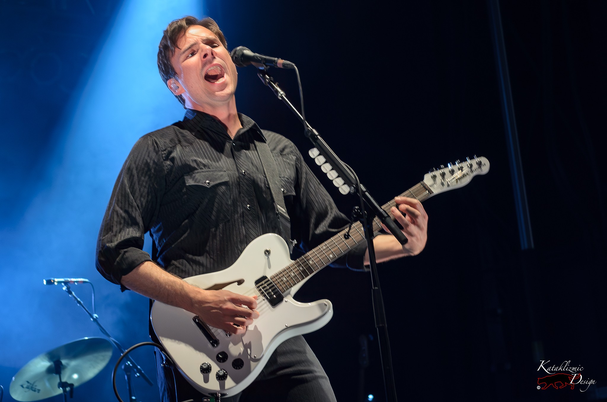 Jim Adkins of Jimmy Eat World performs live on stage with a white electric guitar and black button-down shirt, under blue stage lighting