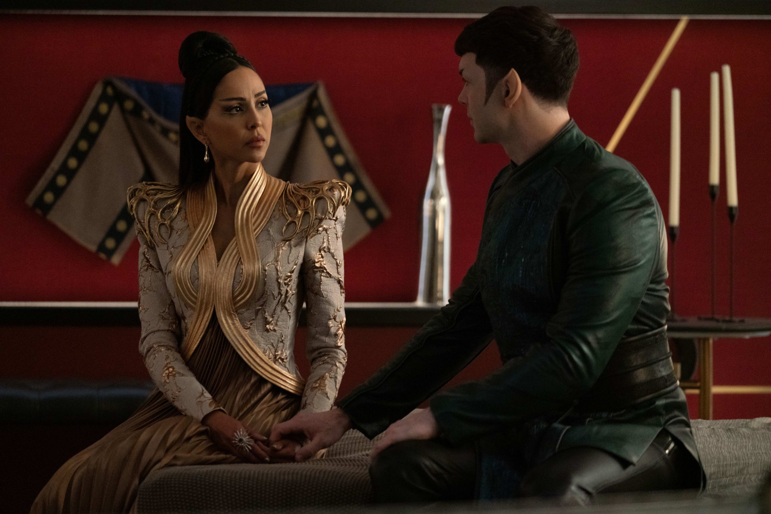 Spock and T’Pring sit close on a bench, holding hands solemnly as she looks at him with disappointment; their ornate Vulcan surroundings underscore the formality and tension of the moment.