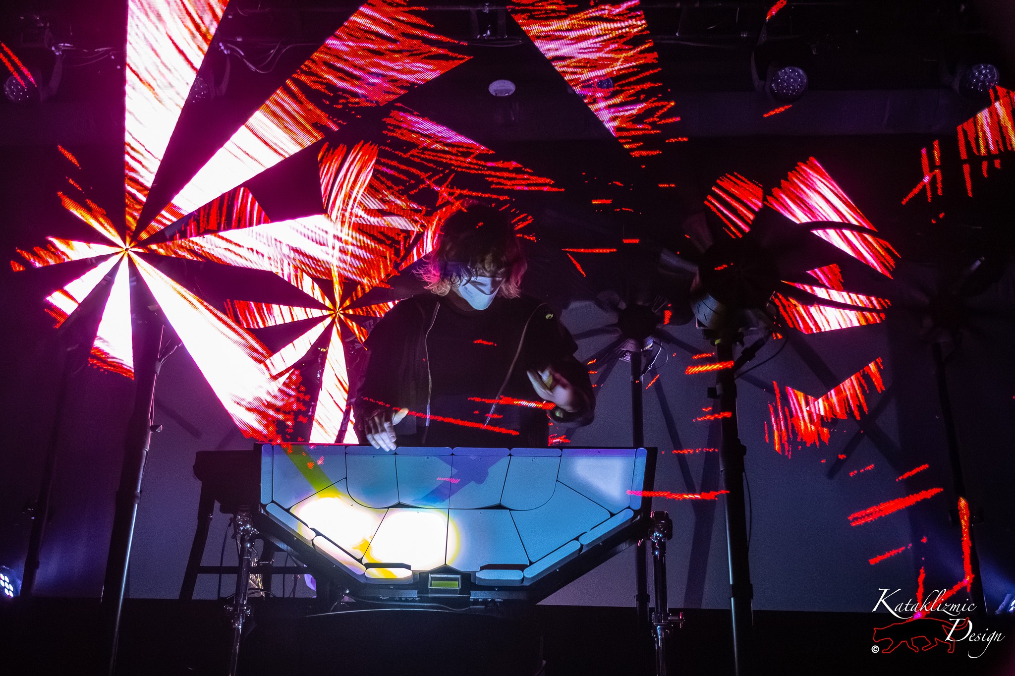 Corin Roddick of Purity Ring performs in a mask behind an illuminated console as red fan-like visuals surround him on stage in Phoenix.