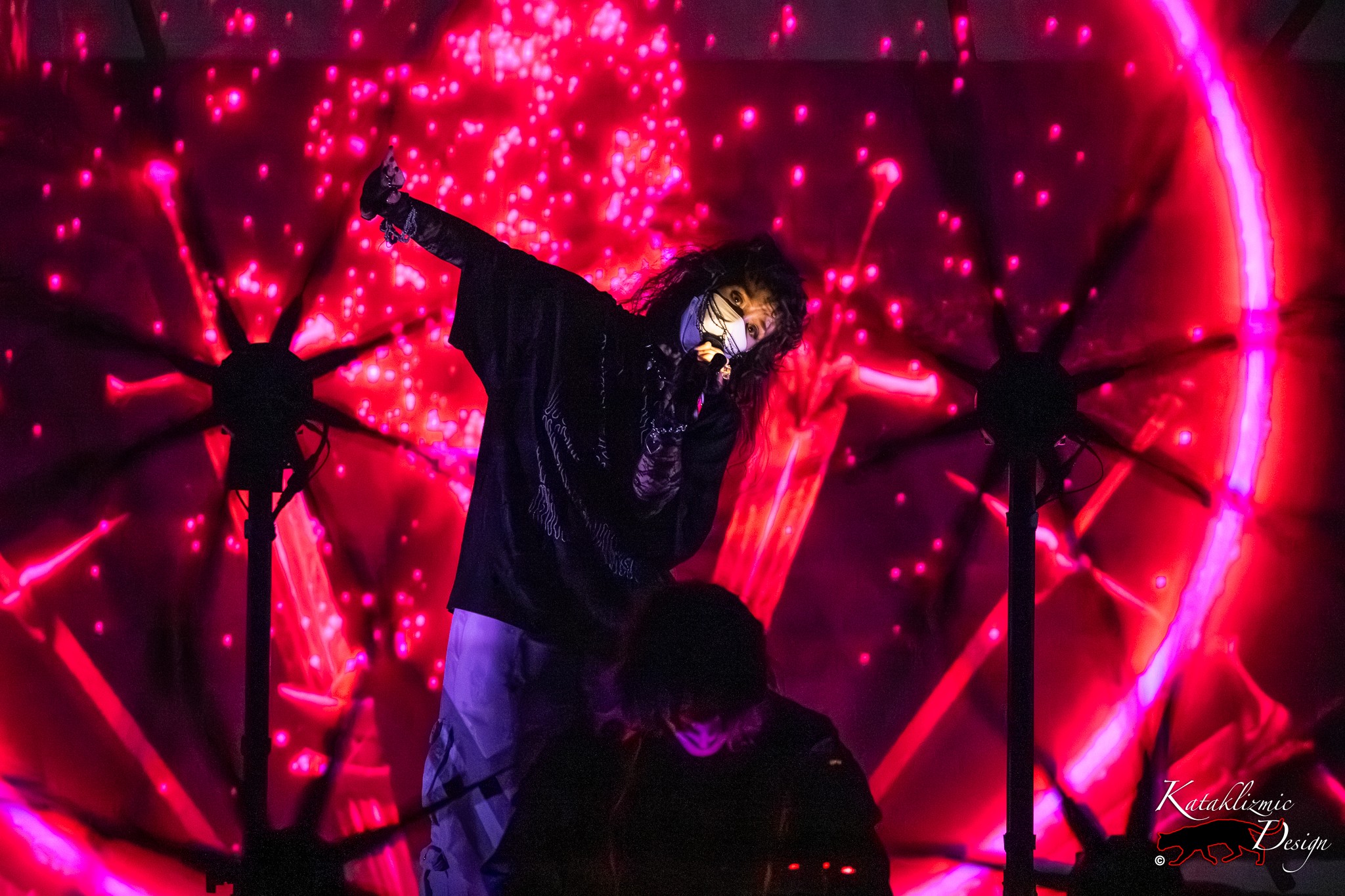 ALT: Megan James of Purity Ring, masked and dressed in black, leans sideways into the mic as vivid red light patterns radiate behind her, with Corin Roddick faintly visible at the console below.