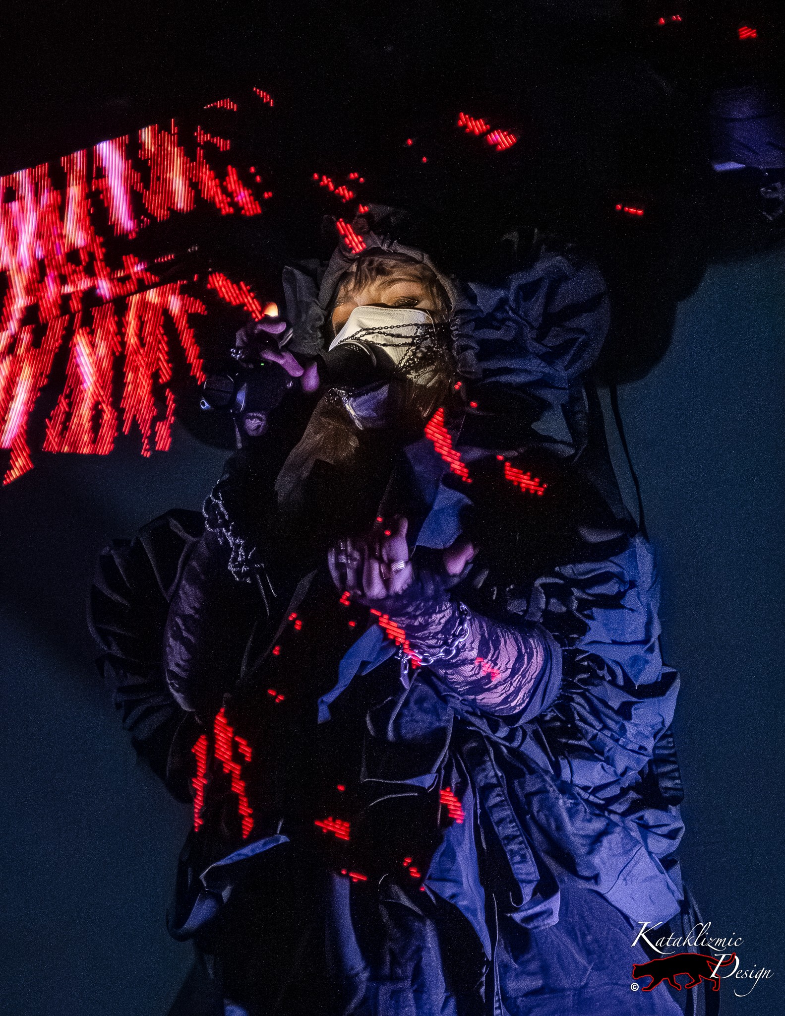 Megan James of Purity Ring sings into a microphone while wrapped in a voluminous dark coat and white mask, scattered red LED streaks cutting across her in the otherwise dark stage at The Van Buren in Phoenix.