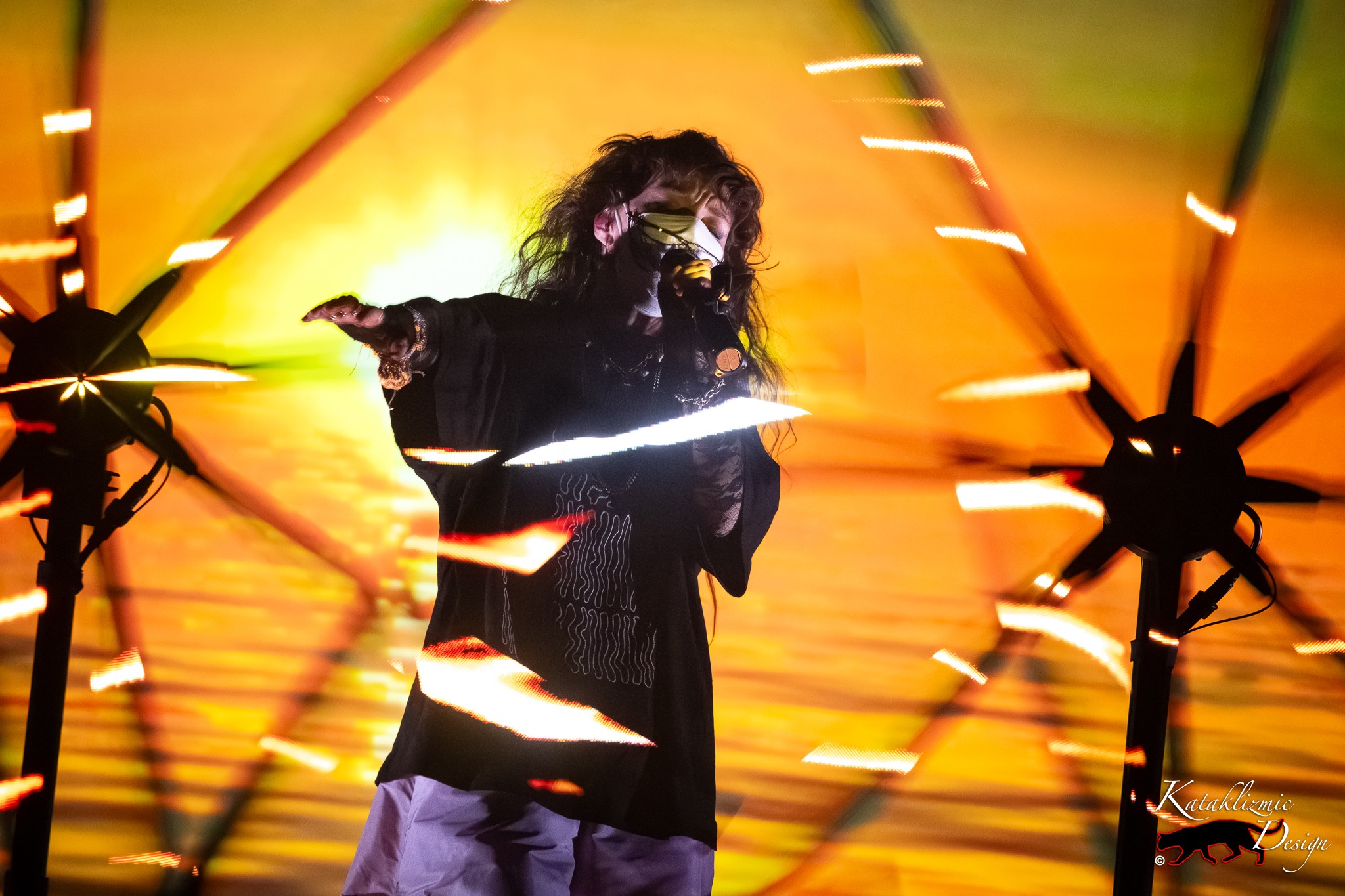 Megan James of Purity Ring sings into the microphone with her arm extended as streaks of golden light spin around her against a vivid orange backdrop.