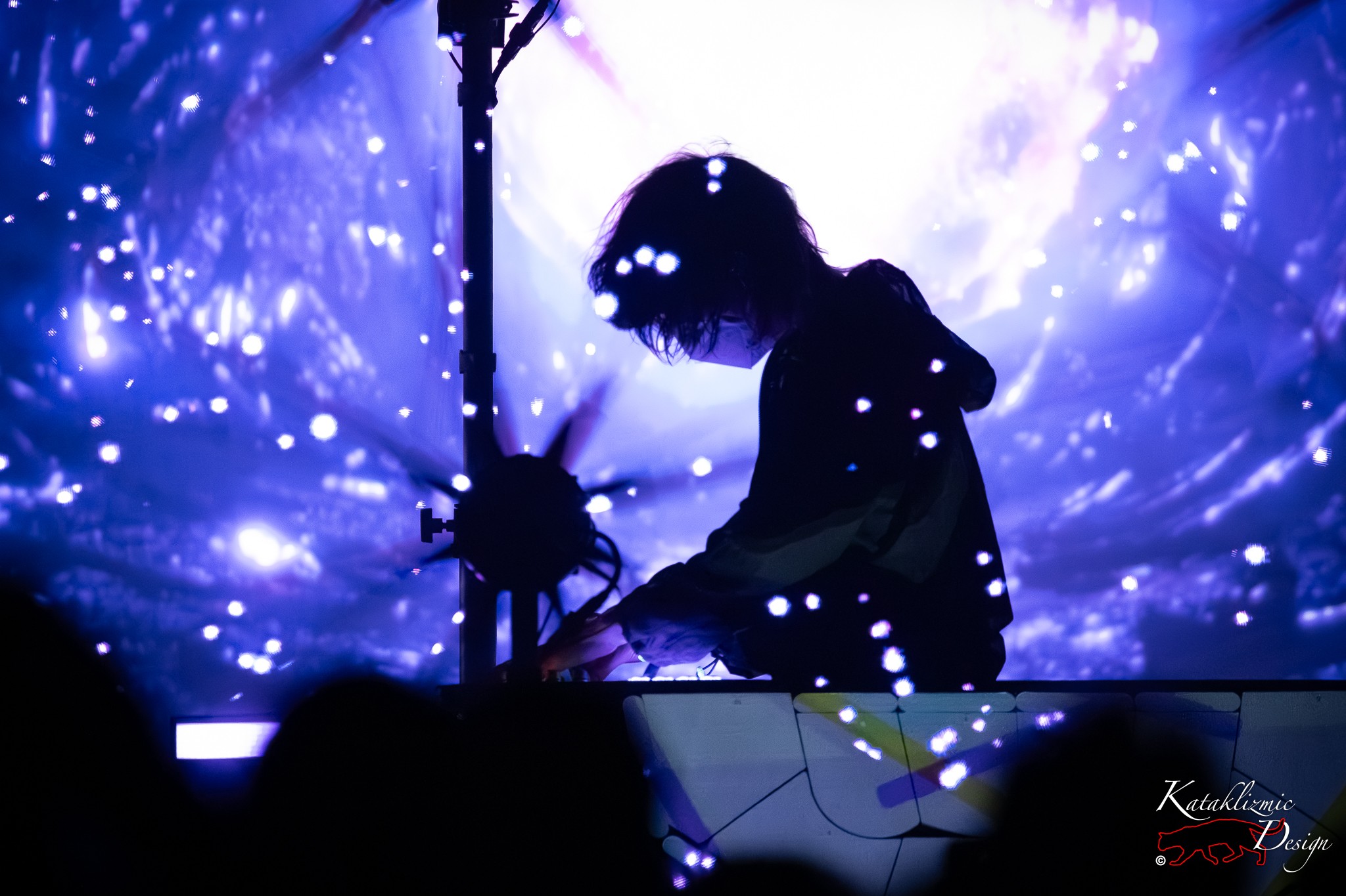 Purity Ring producer Corin Roddick silhouetted at his console as blue galaxy-like visuals and sparkling light particles swirl across the stage.