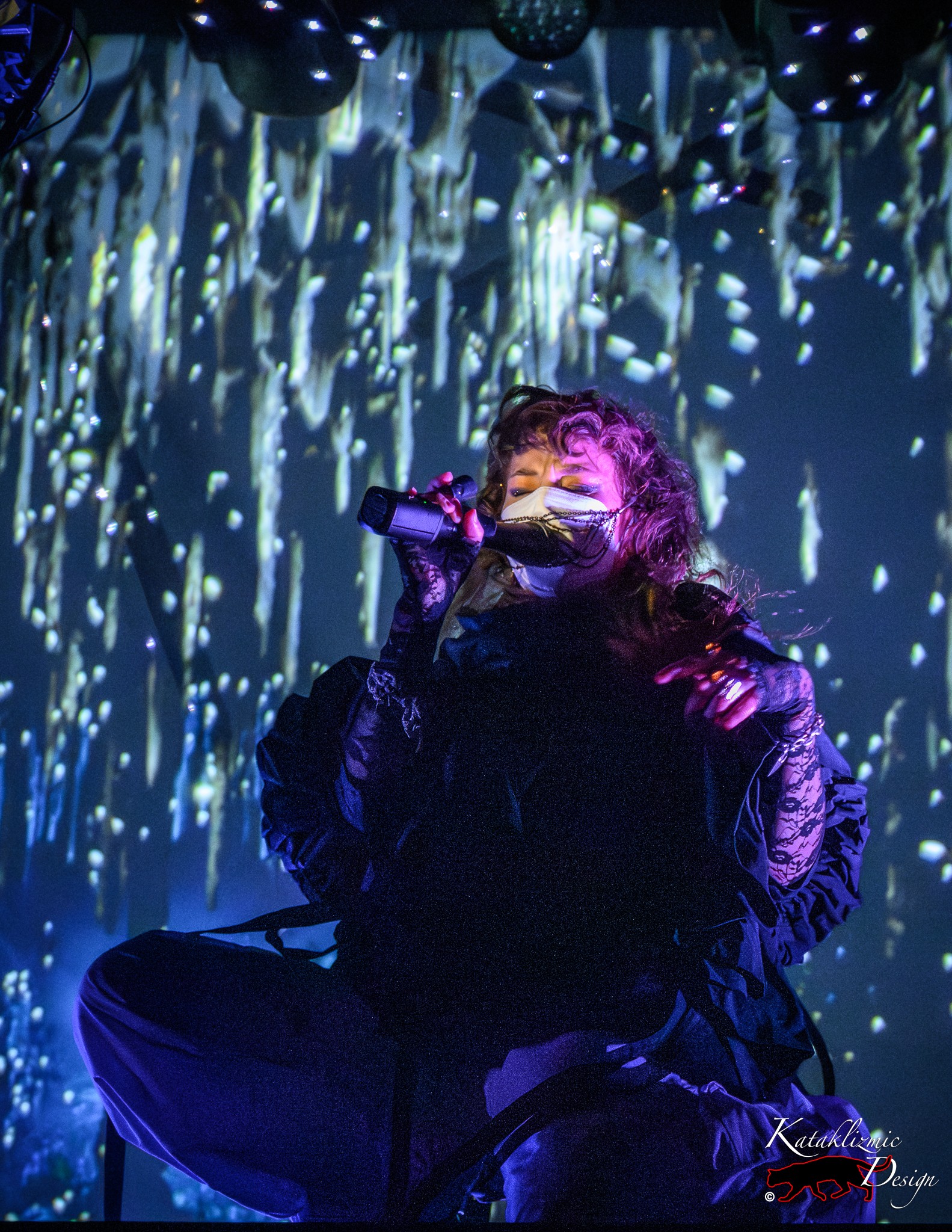 Megan James of Purity Ring sits cross-legged onstage, masked and dressed in black, singing into a handheld mic as cool blue and white light streaks fall in the background.