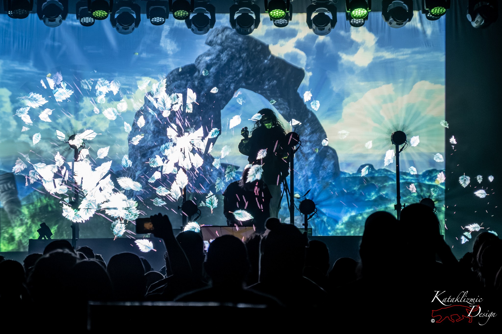 Purity Ring perform in silhouette as bright holographic leaf shapes burst from a giant blue seascape screen, drifting toward the crowd in the foreground.