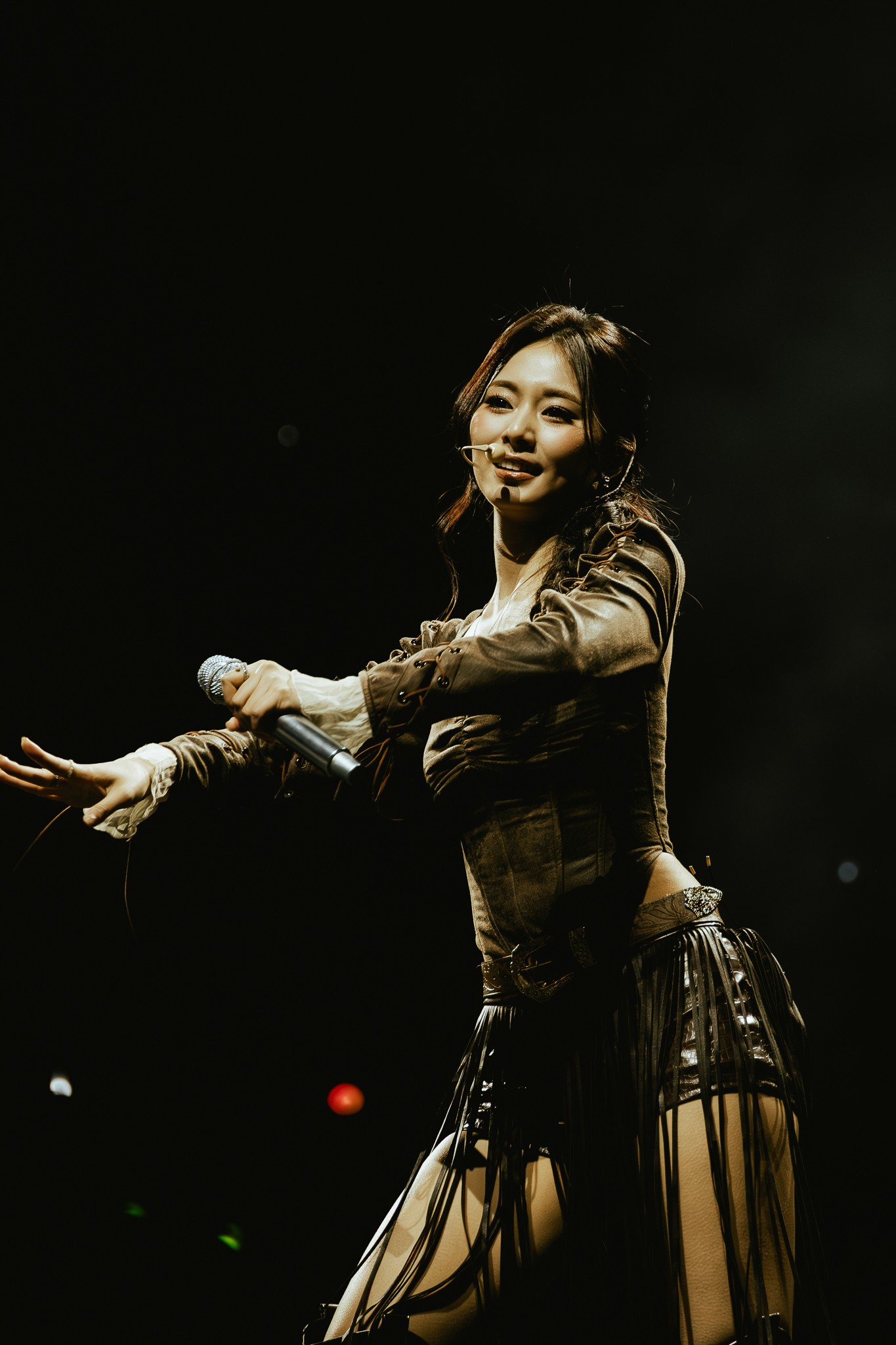 ALT text (plain):
Tzuyu of TWICE performs onstage holding a microphone, reaching outward under warm spotlighting against a darkened arena backdrop.