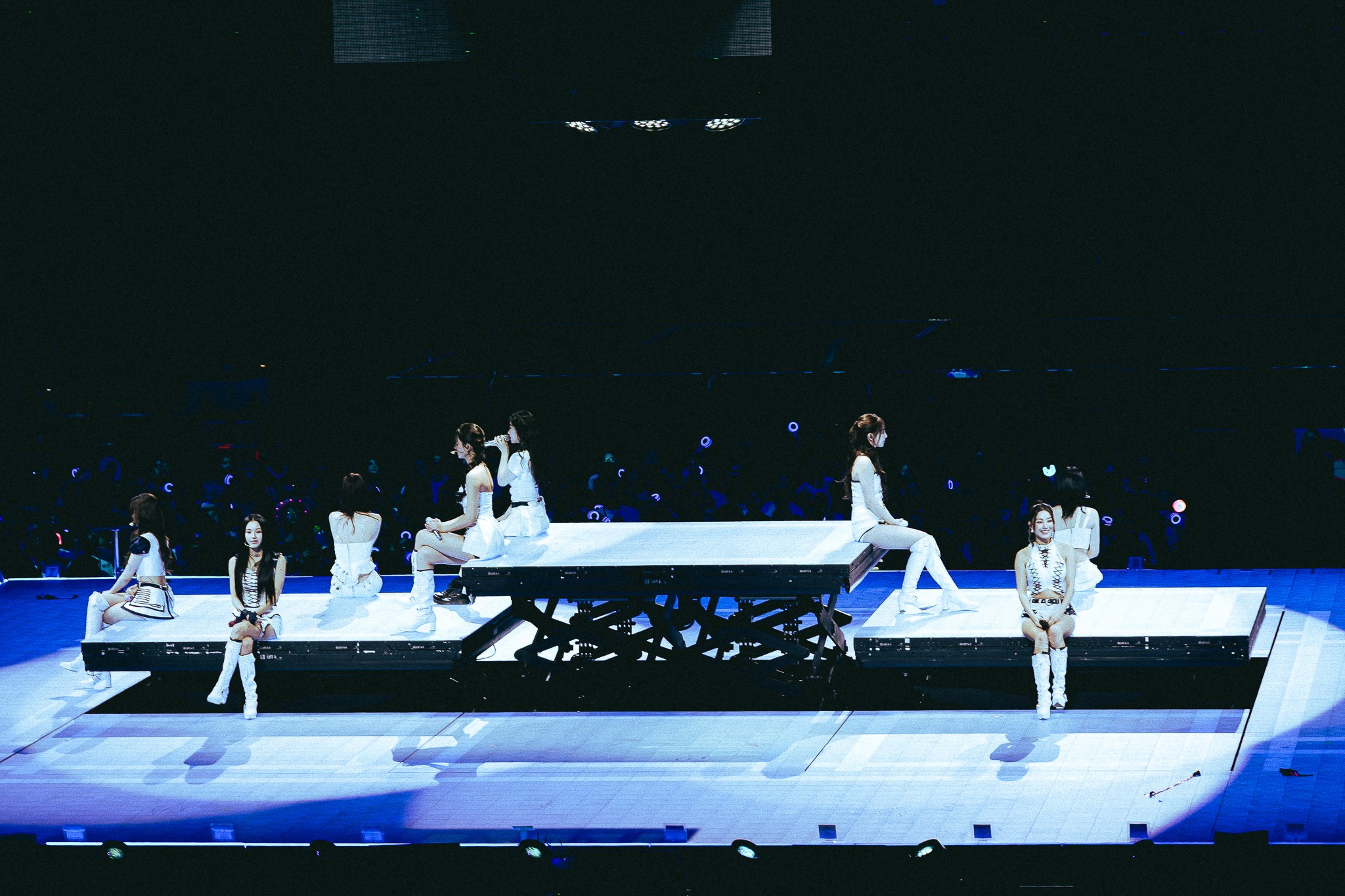 Members of TWICE sit across a tiered stage in coordinated white outfits, illuminated by blue lighting as the audience's light sticks glow in the background.
