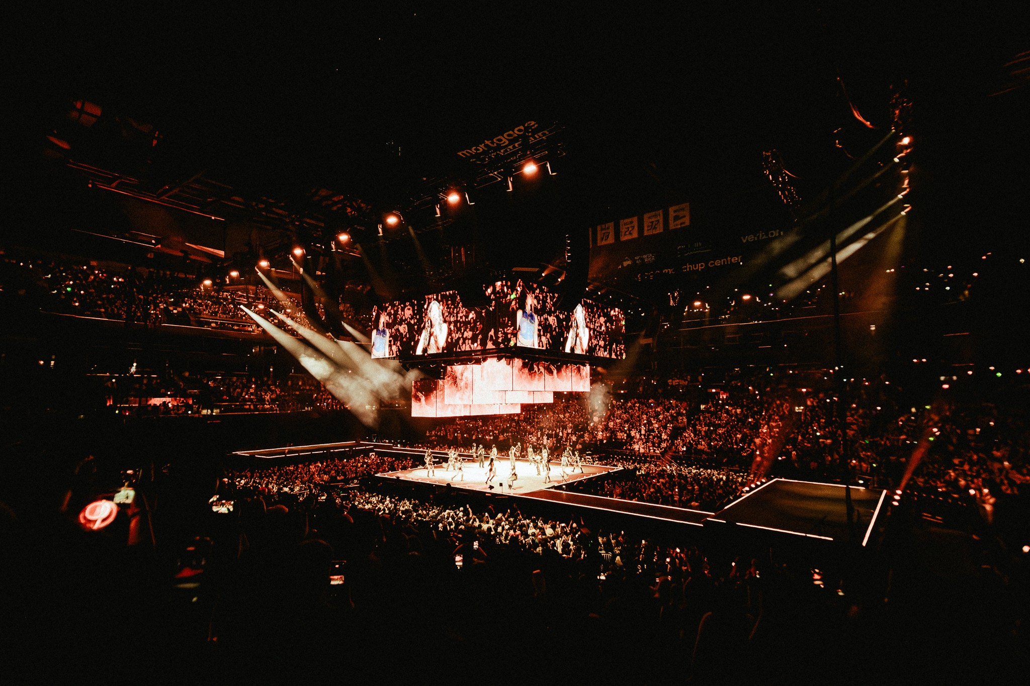 TWICE performs on a central stage inside a packed arena, with large overhead screens displaying the group as warm amber lighting spreads across the audience.
