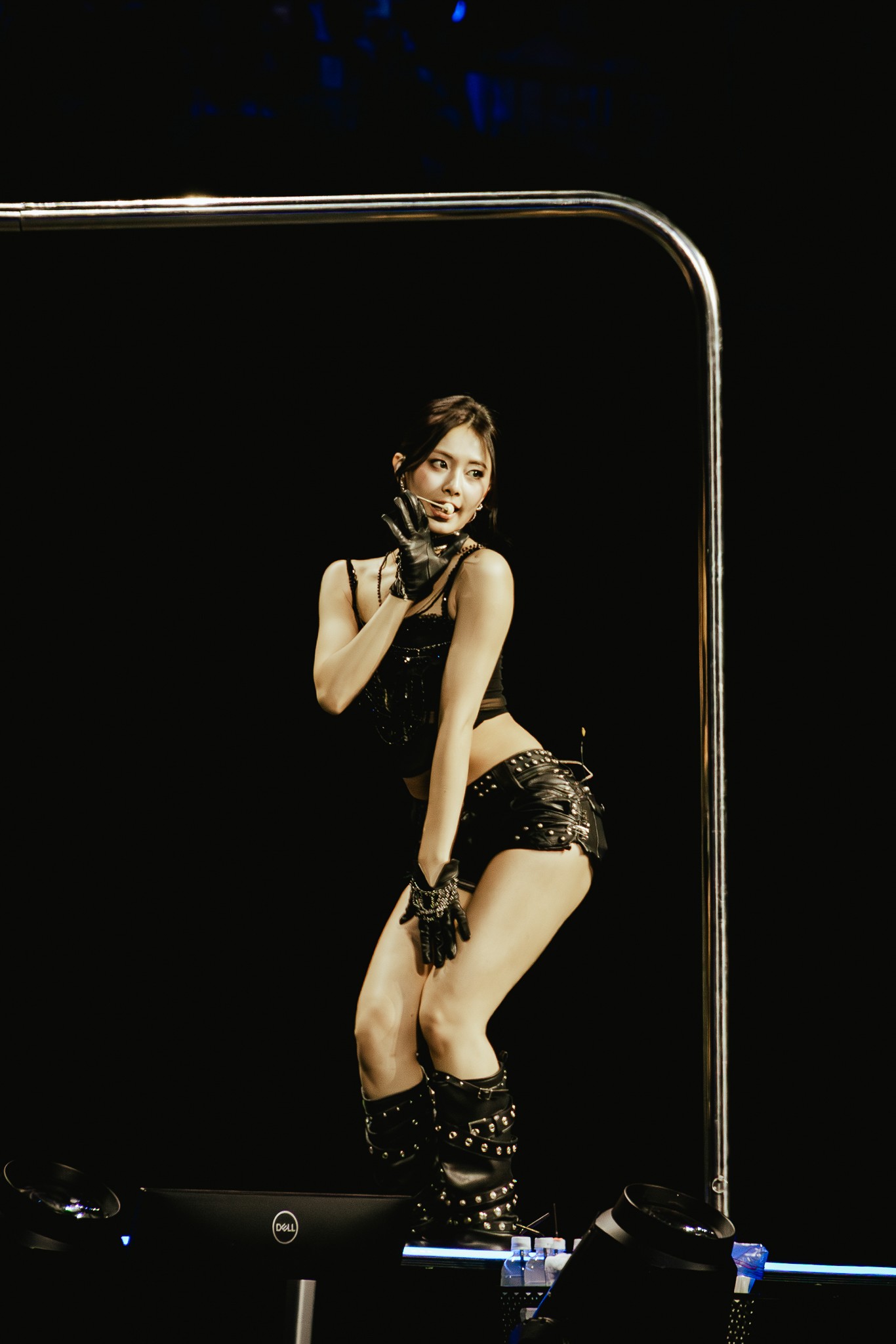 Tzuyu of TWICE performs her solo beside a metal prop, posed in black stagewear under low blue lighting.