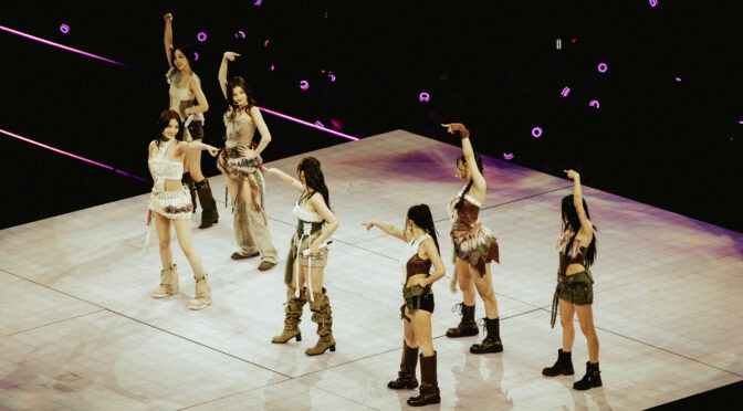 TWICE stands together on a raised stage, arms lifted in a closing formation as purple fan lightsticks glow throughout the arena.