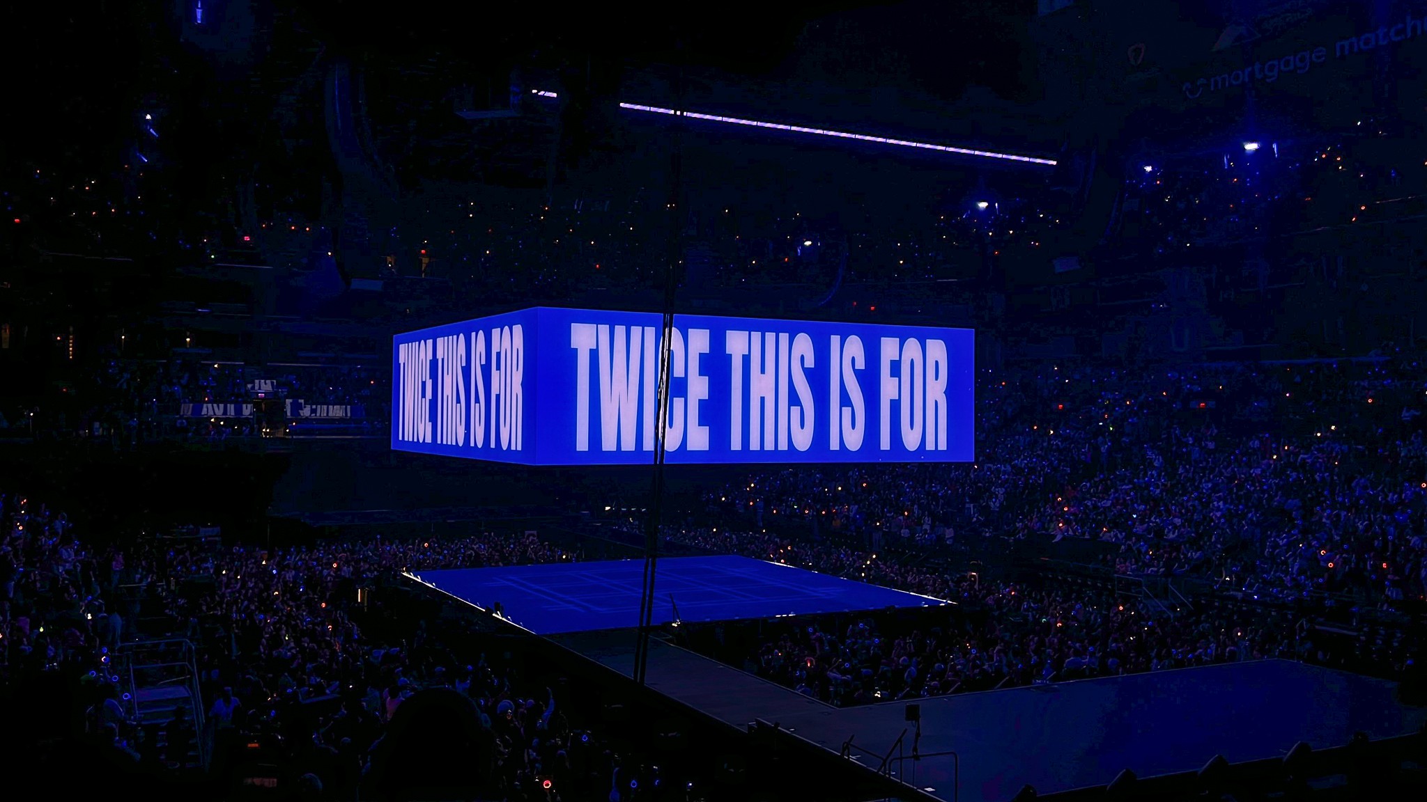 A wide view of a packed arena during TWICE’s “THIS IS FOR” World Tour, with a large blue overhead screen displaying the words “TWICE THIS IS FOR” above the main stage as fans hold glowing light sticks throughout the crowd.