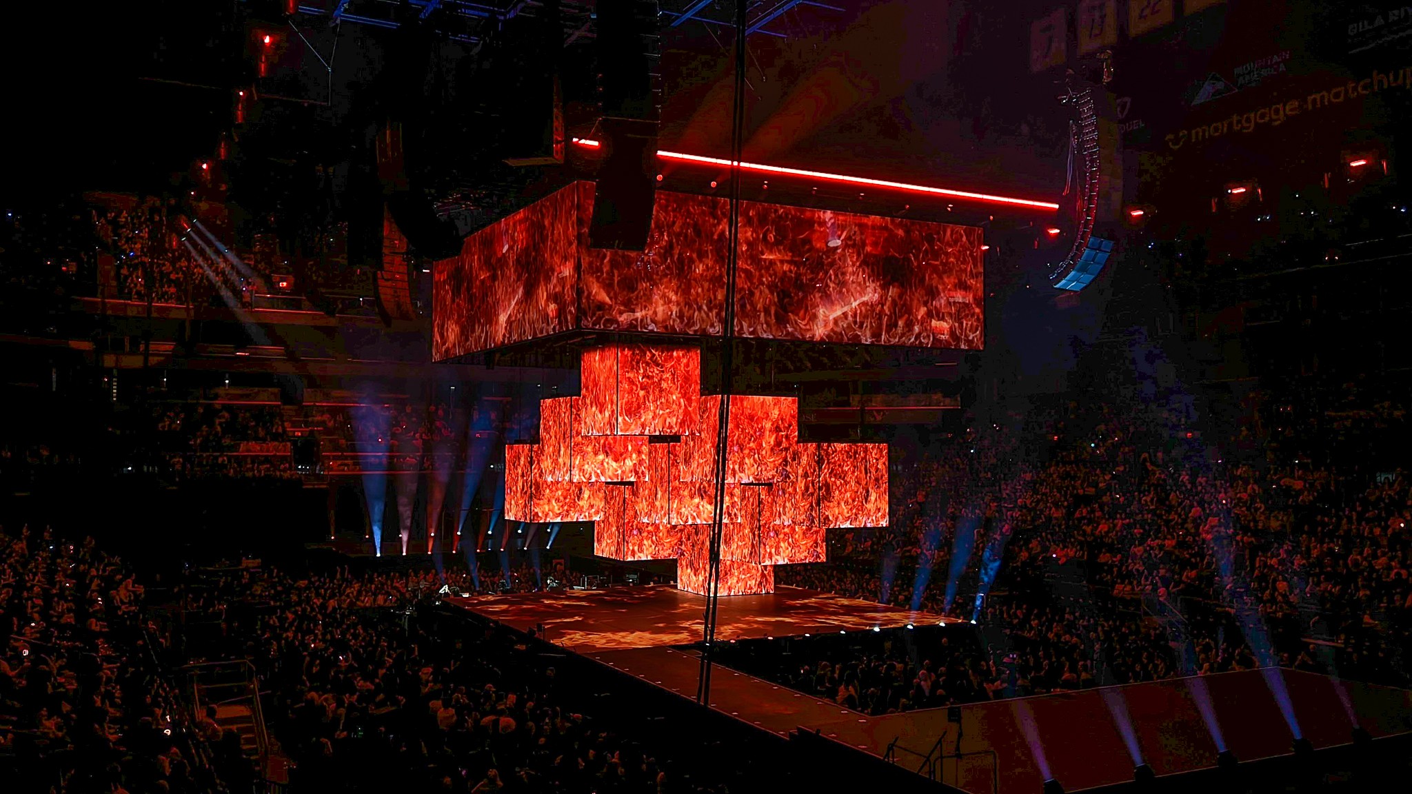 A wide view of the arena shows a large, multi-tiered screen glowing red above the stage as sections lower toward the floor, with a packed audience surrounding the performance area.