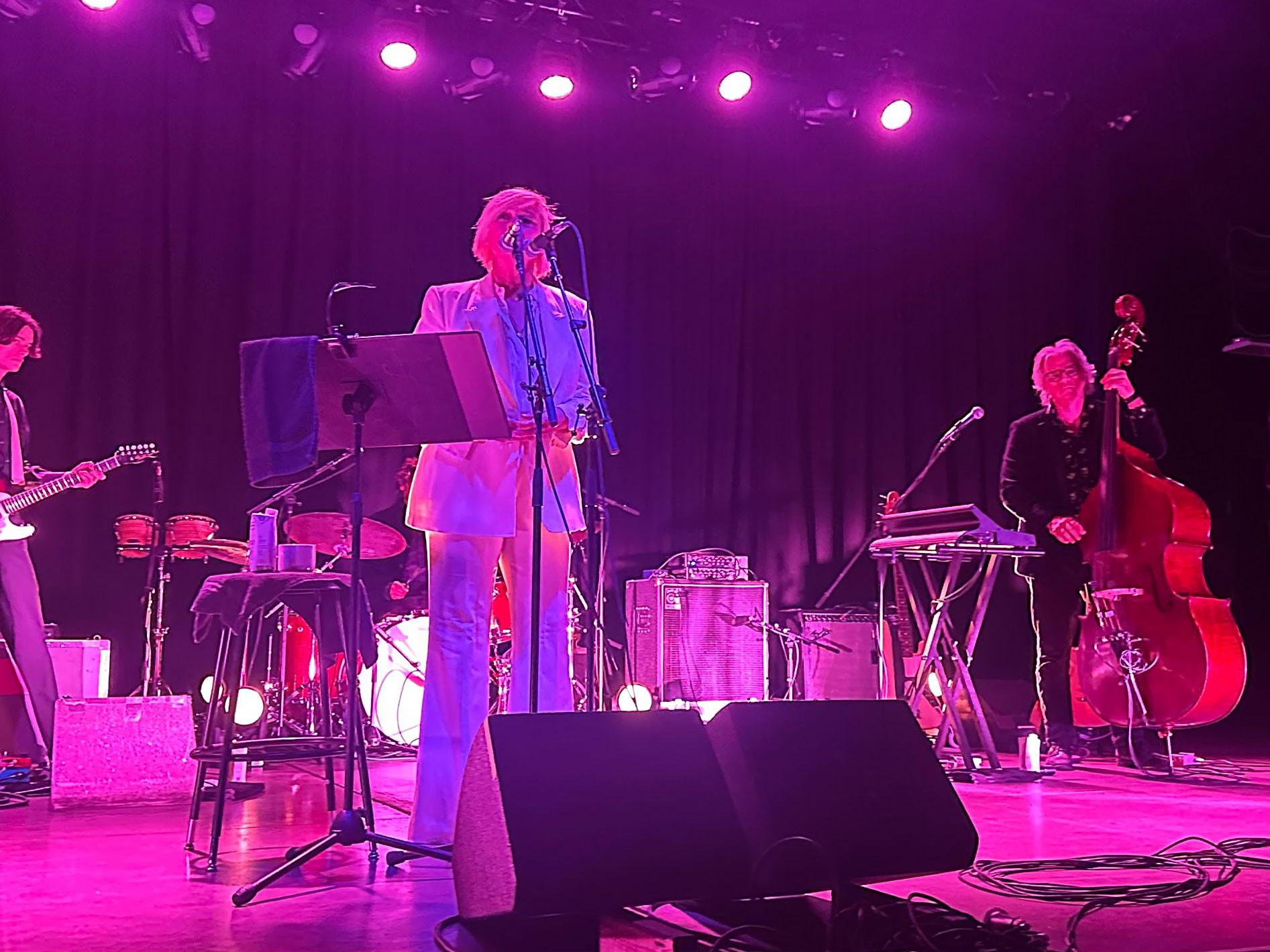 Cat Power performs at The Van Buren in Phoenix, standing at a microphone in a bright white suit under vivid magenta stage lights. A guitarist, drummer, and upright bassist are visible behind her, with stage monitors and cables in the foreground.