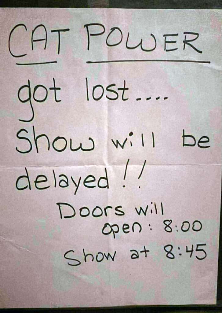 A handwritten sign on white paper reads: “CAT POWER got lost… Show will be delayed!! Doors will open: 8:00. Show at 8:45.” The paper appears slightly wrinkled and was posted at the venue entrance to inform concertgoers of the delay.