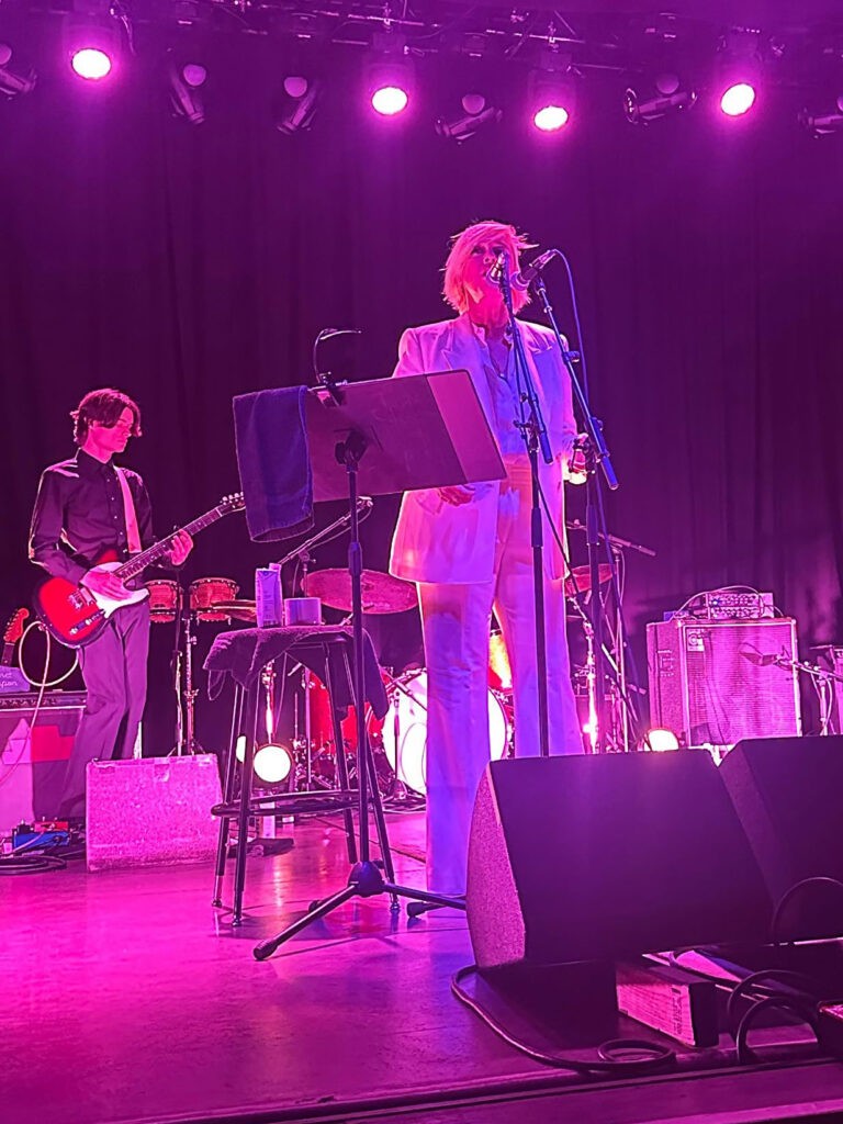 Cat Power stands at a microphone at The Van Buren in Phoenix, wearing a bright white suit under vivid magenta stage lighting. A guitarist and drum kit are visible behind her, with stage monitors and equipment in the foreground.