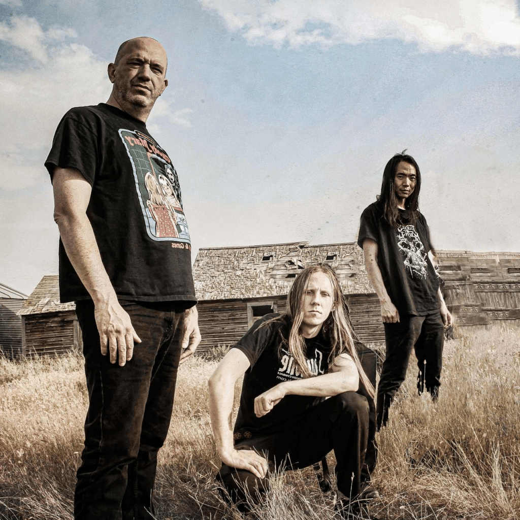 Press photo of extreme metal band Narcotic Wasteland standing in a field in front of a weathered wooden structure. Three band members pose with a gritty, confrontational presence under an open sky, wearing black metal shirts, reflecting the band’s raw, aggressive aesthetic and modern extreme metal identity.