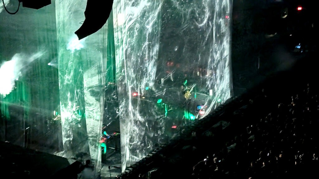 Side-angle view of the stage draped in translucent curtains, with green lighting and swirling black-and-white projections partially revealing the band while the crowd watches in the dark.