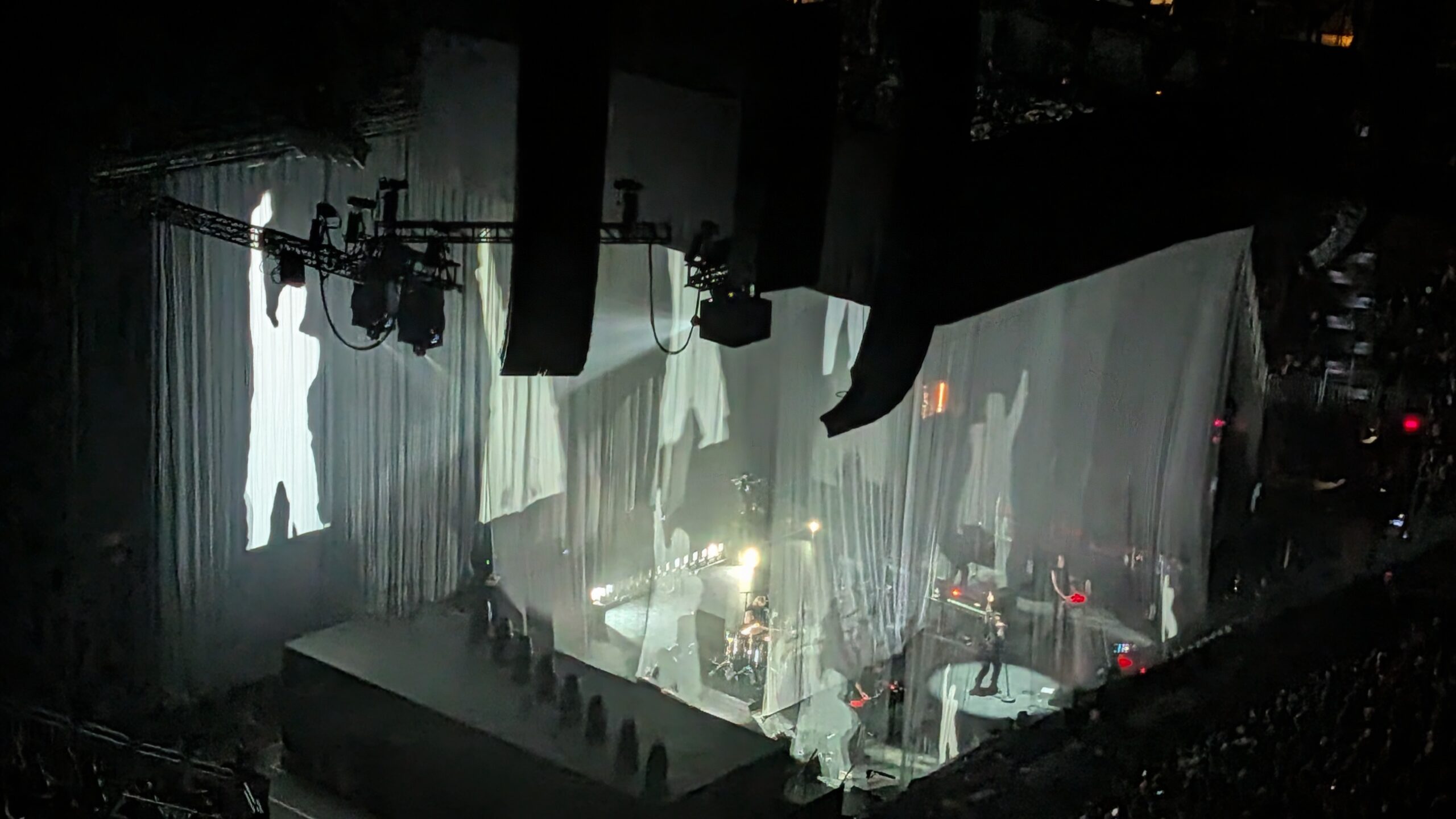 Wide arena view of the band playing behind translucent drapes, with bright backlighting projecting large silhouettes onto the curtains and stage lighting rigs hanging overhead.