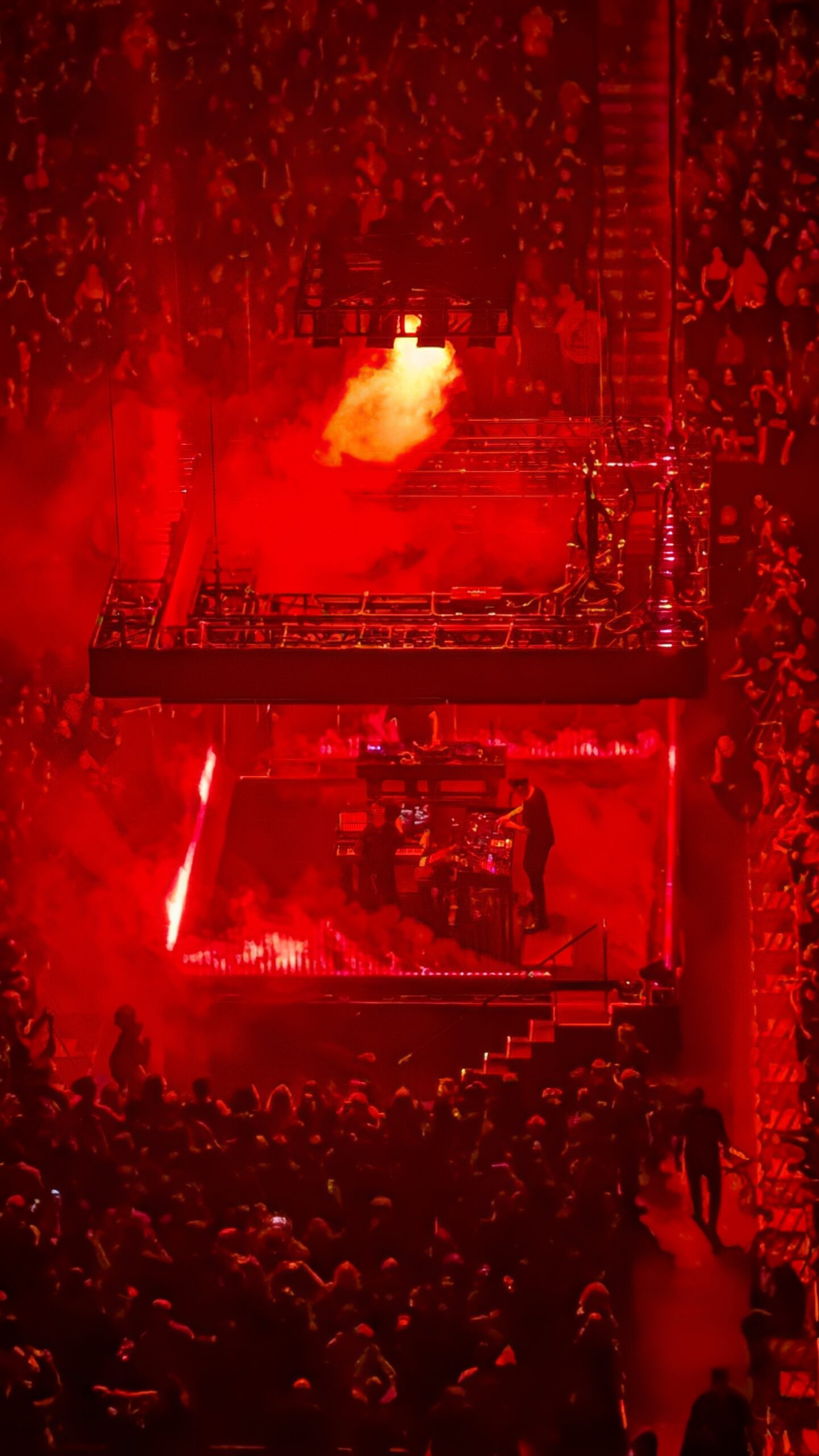 High-angle view of Nine Inch Nails performing on a center platform under intense red lighting and smoke, with a suspended lighting rig overhead and a packed arena crowd surrounding the stage in Glendale, Arizona.
