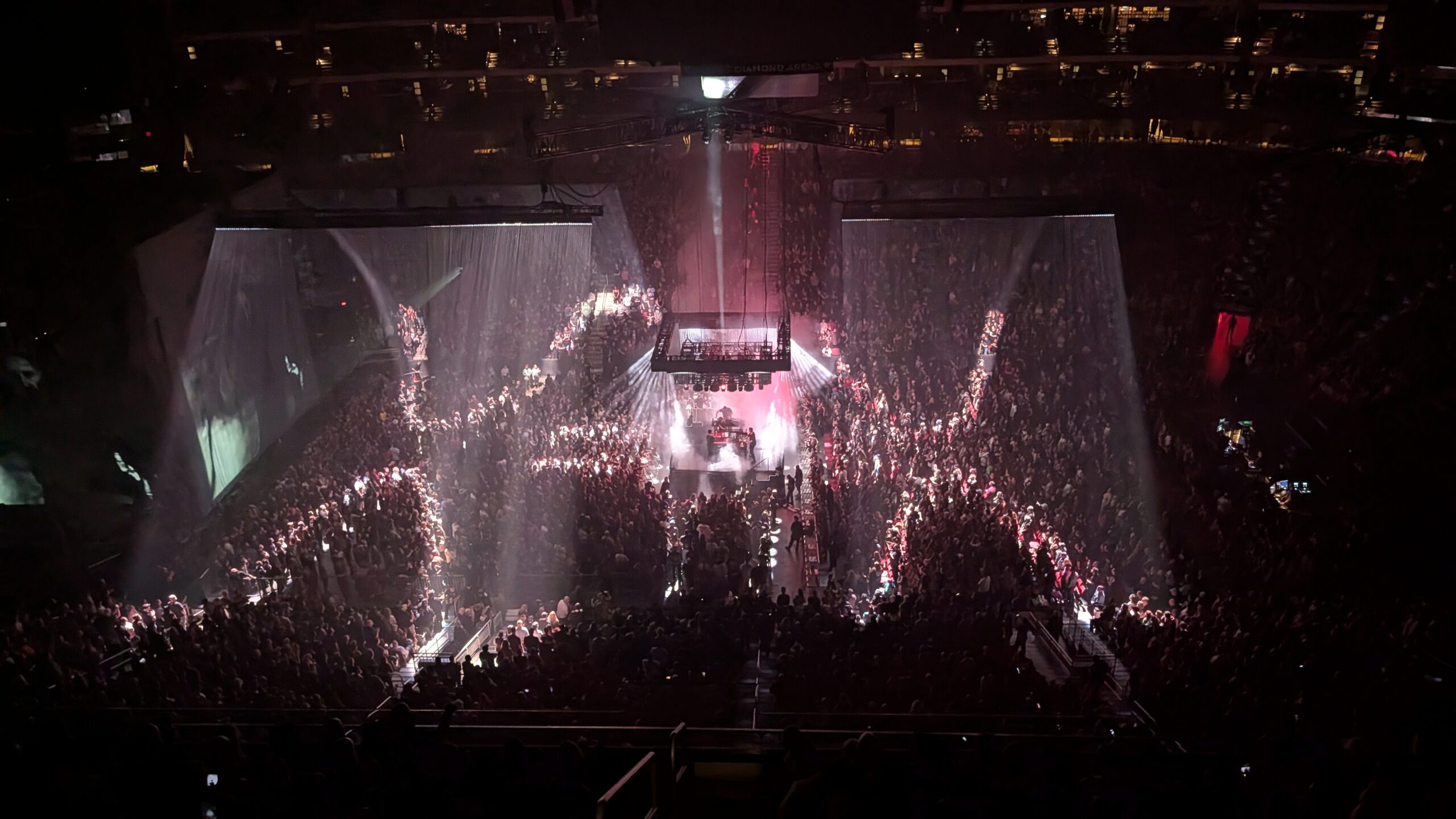 High, wide shot of a packed arena with a small center stage surrounded by the crowd, as bright white beams cut through haze and cast rippling, curtain-like light patterns across sections of the audience.