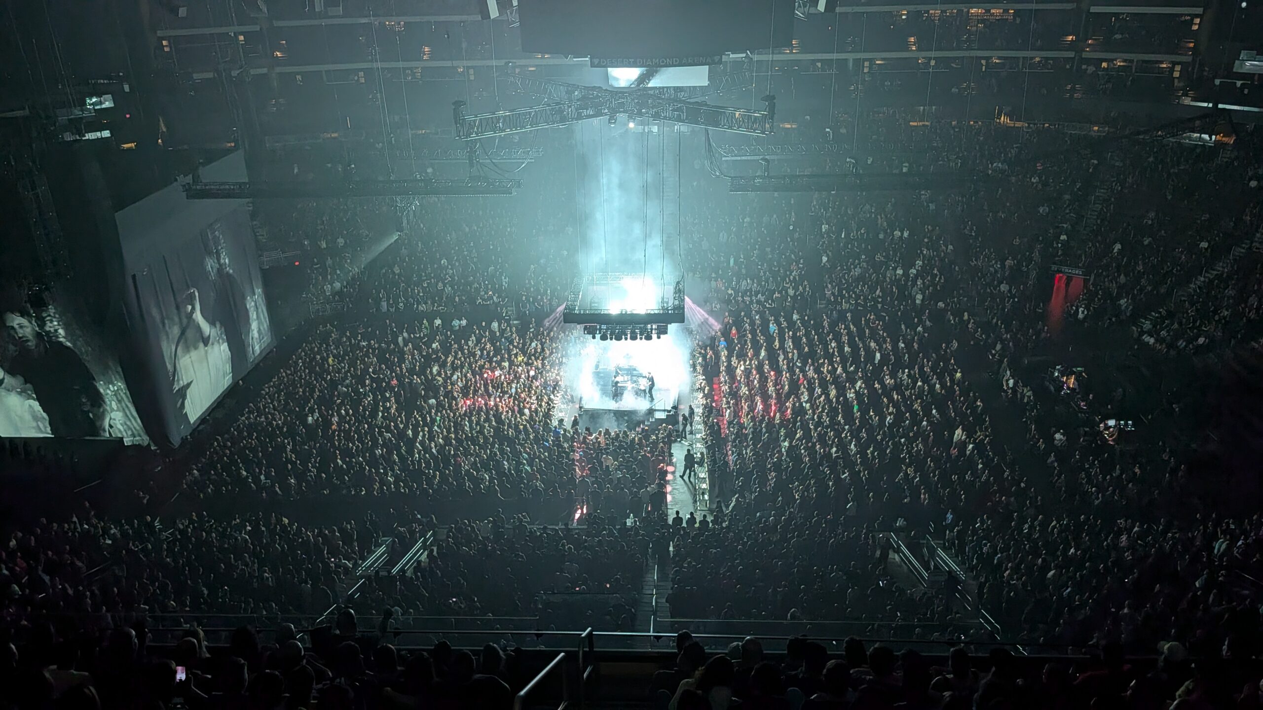 High, wide arena view with a bright white glow and haze rising from the center stage, surrounded by a dense crowd, with a large side screen showing black-and-white live footage.