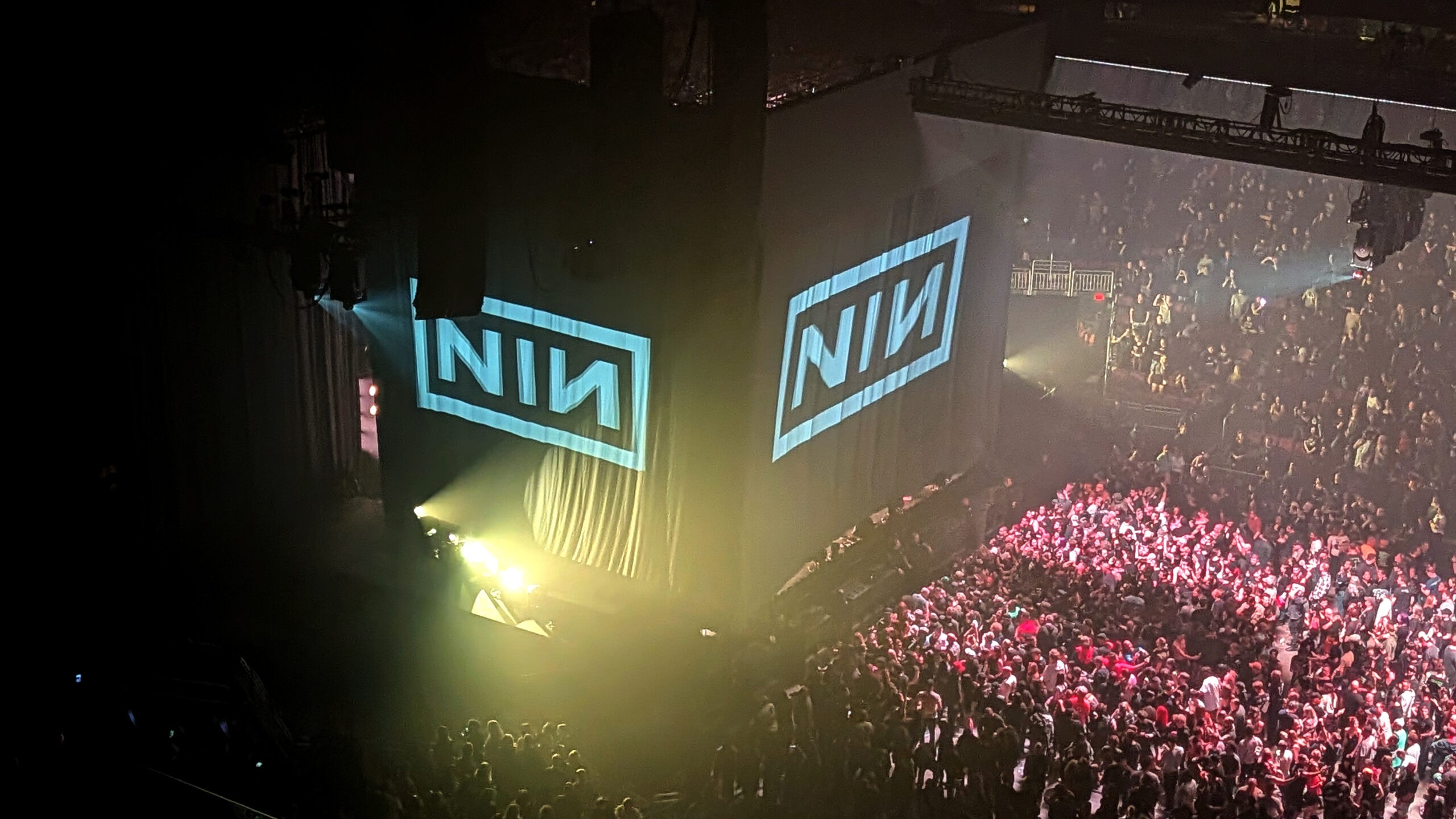 High-angle view of the arena after the concert, with the NIN logo projected on large hanging curtains as the crowd gathers and begins to clear the floor under hazy yellow light.
