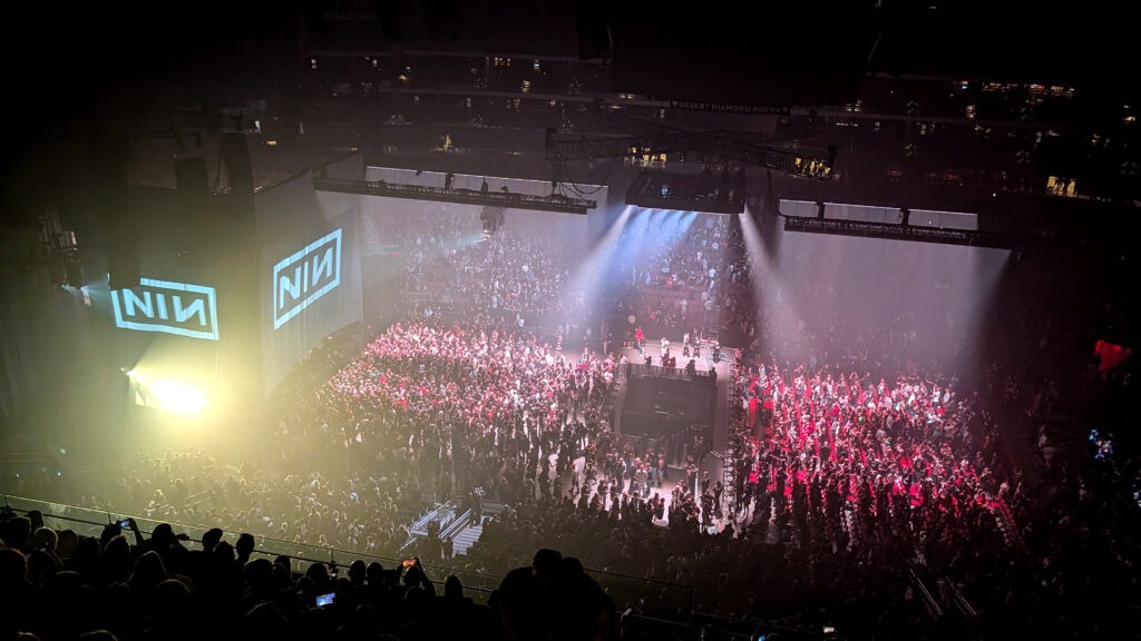 Wide crowd shot inside an arena during a Nine Inch Nails concert, with the NIN logo projected on large side screens, bright stage lighting cutting through haze, and a packed floor and seating bowl.