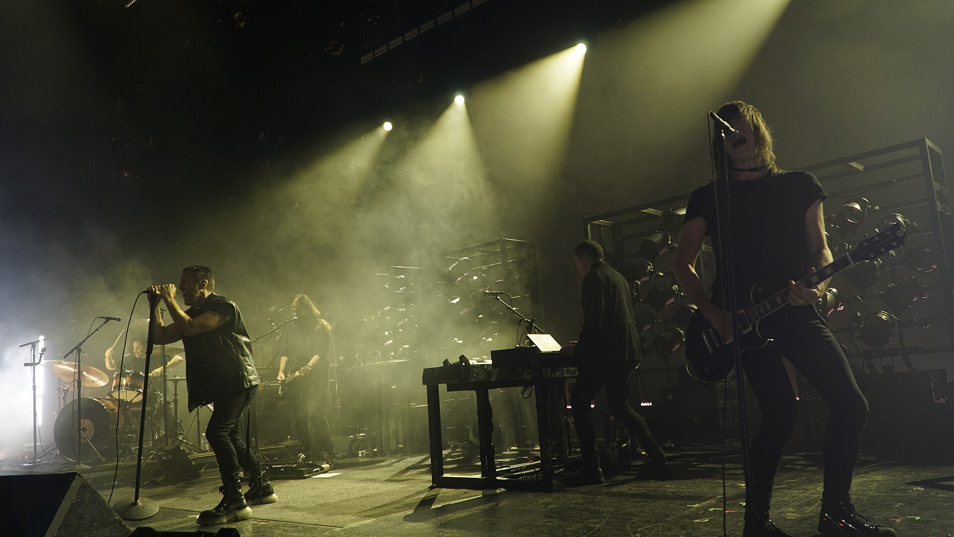 Wide stage photo of Nine Inch Nails performing live in smoky yellow light, with the vocalist at a microphone, guitarist in the foreground, drummer at left, and lighting rigs behind the band.