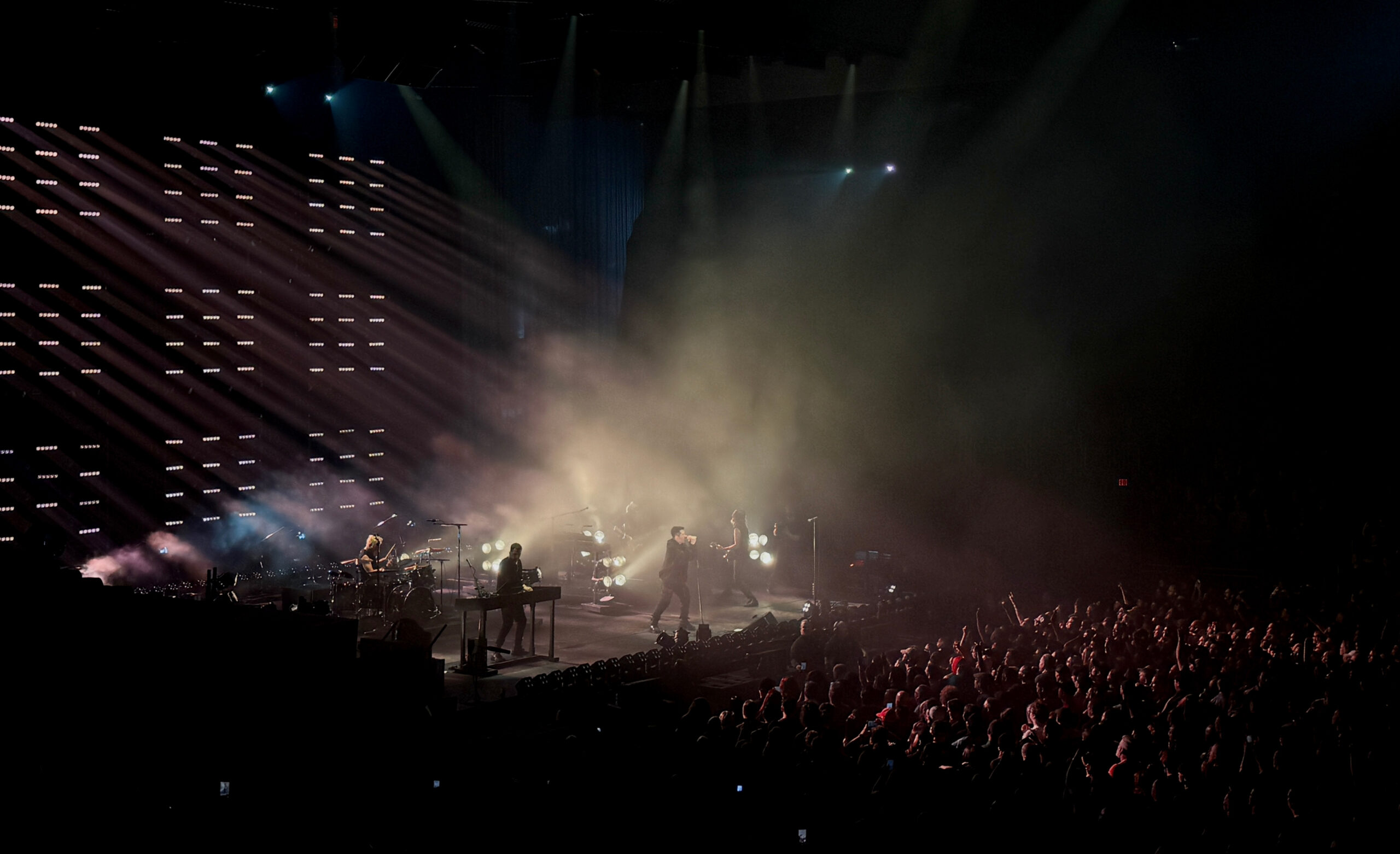 Wide stage view with the band performing amid thick fog, diagonal light beams cutting across a wall of small lights, and a packed audience filling the foreground.