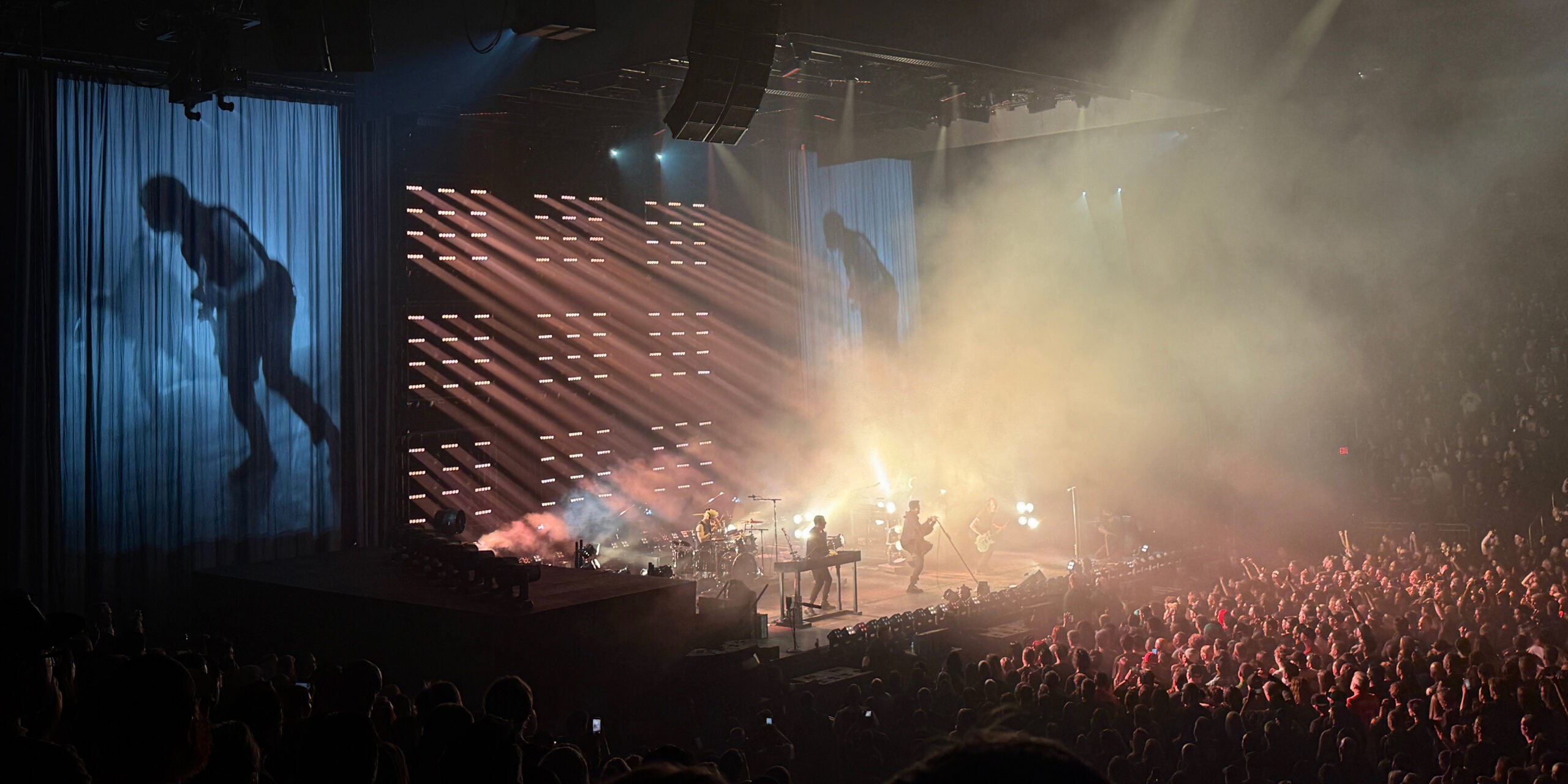 Wide arena view after the sheer curtain lifts, with the full band visible through heavy haze and bright stage lights, while a large silhouette projection looms on a side curtain and the crowd cheers at the front rail.