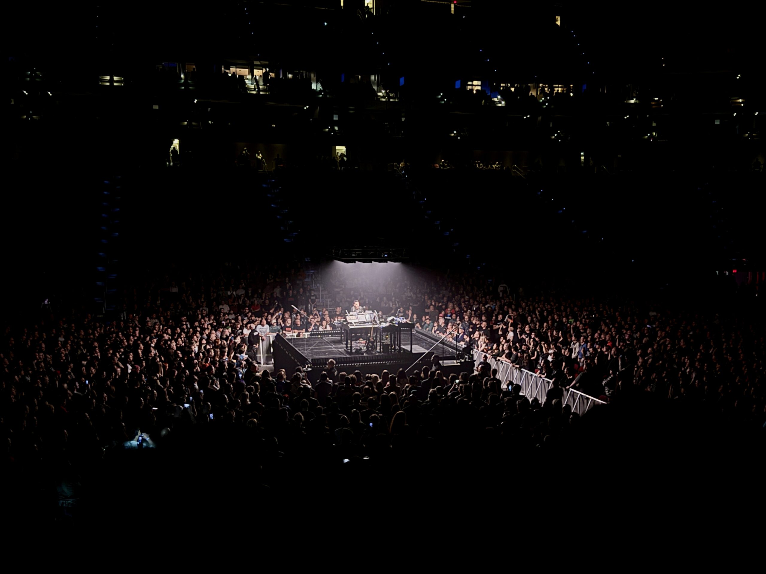 Wide arena shot of Trent Reznor at a small center stage lit by a single white glow, surrounded by a dense crowd in near darkness.