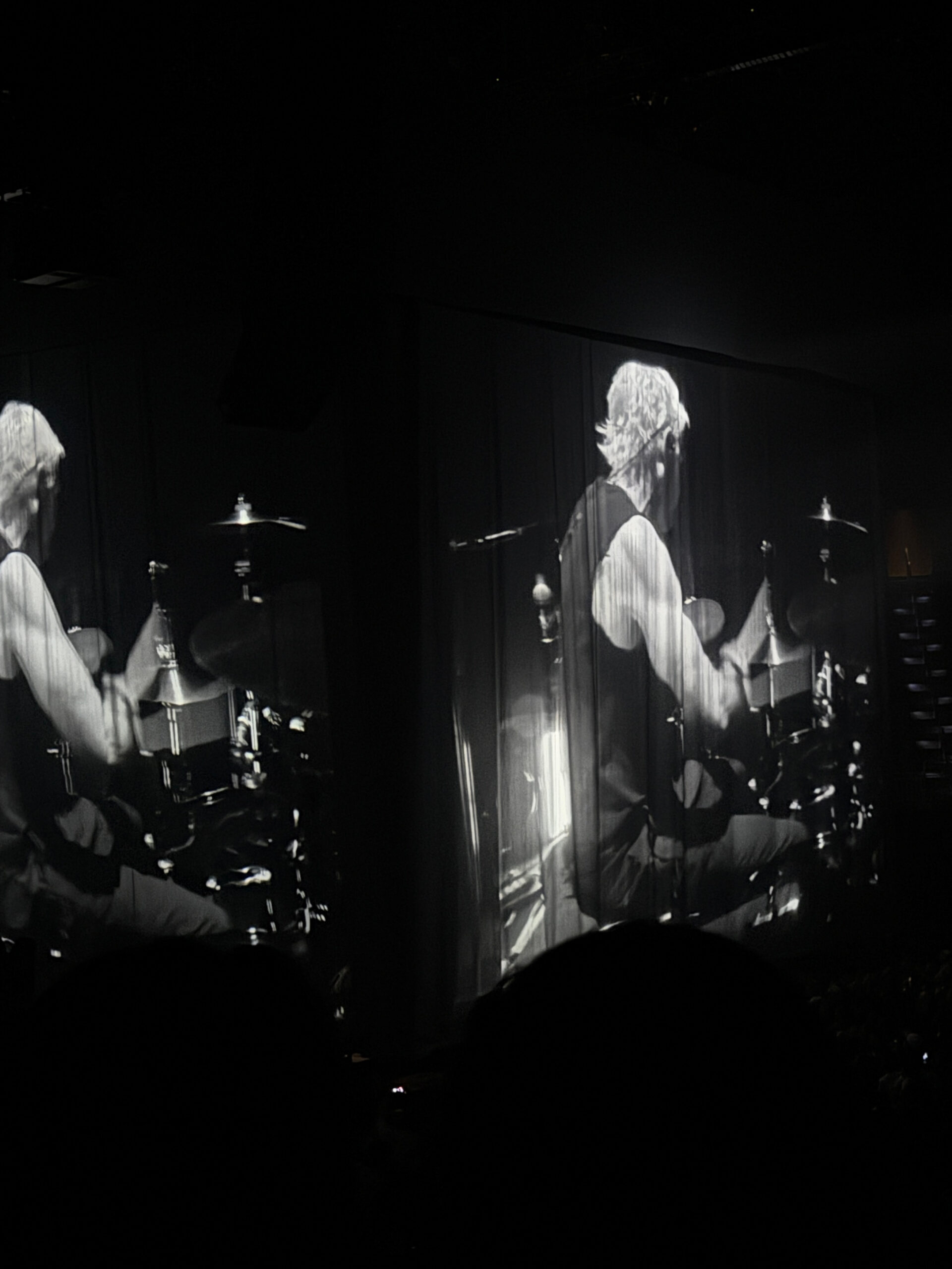 Black-and-white projection of a drummer playing fills large hanging curtains, with the arena around it mostly dark and silhouetted.
