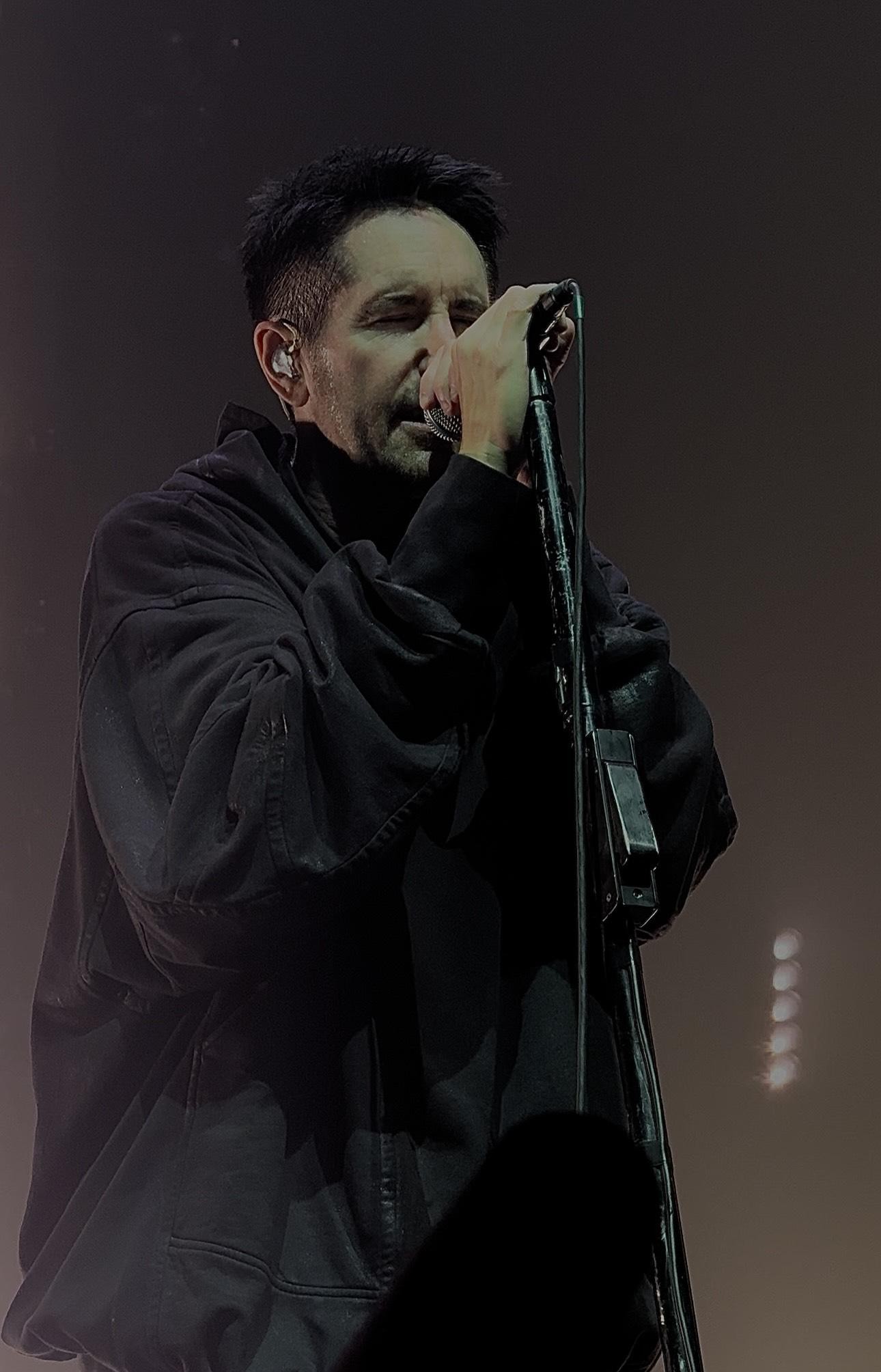 Close-up concert photo of Trent Reznor singing into a microphone on a dim, smoky stage, wearing a dark oversized jacket with in-ear monitors visible.