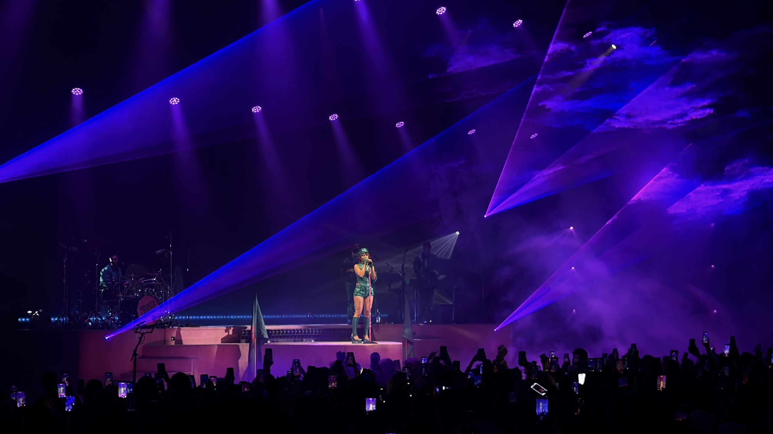 Wide audience photo of Mariah the Scientist performing on the Hearts Sold Separately Tour at Arizona Financial Theatre in Phoenix, standing alone under purple lasers and haze while the crowd watches and records.