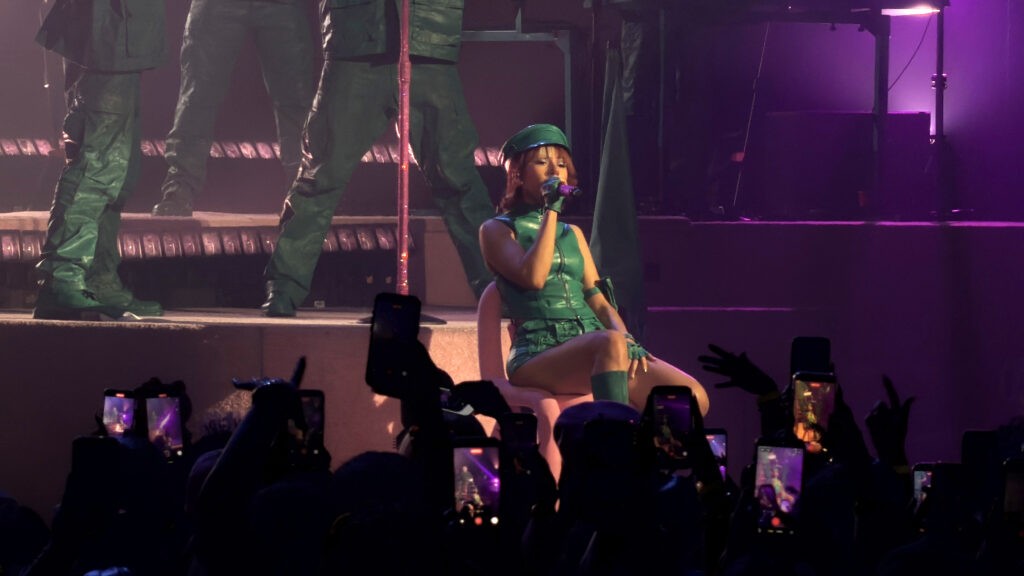 Mariah the Scientist performs seated onstage in a green outfit during the “Hearts Sold Separately Tour” at Arizona Financial Theatre in Phoenix, with fans filming and purple stage lighting filling the venue.