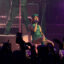 Mariah the Scientist performs seated onstage in a green outfit during the “Hearts Sold Separately Tour” at Arizona Financial Theatre in Phoenix, with fans filming and purple stage lighting filling the venue.