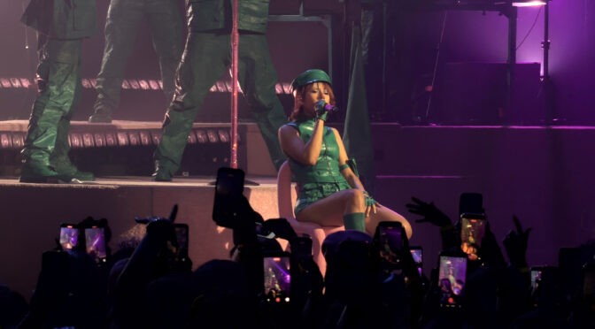 Mariah the Scientist performs seated onstage in a green outfit during the “Hearts Sold Separately Tour” at Arizona Financial Theatre in Phoenix, with fans filming and purple stage lighting filling the venue.
