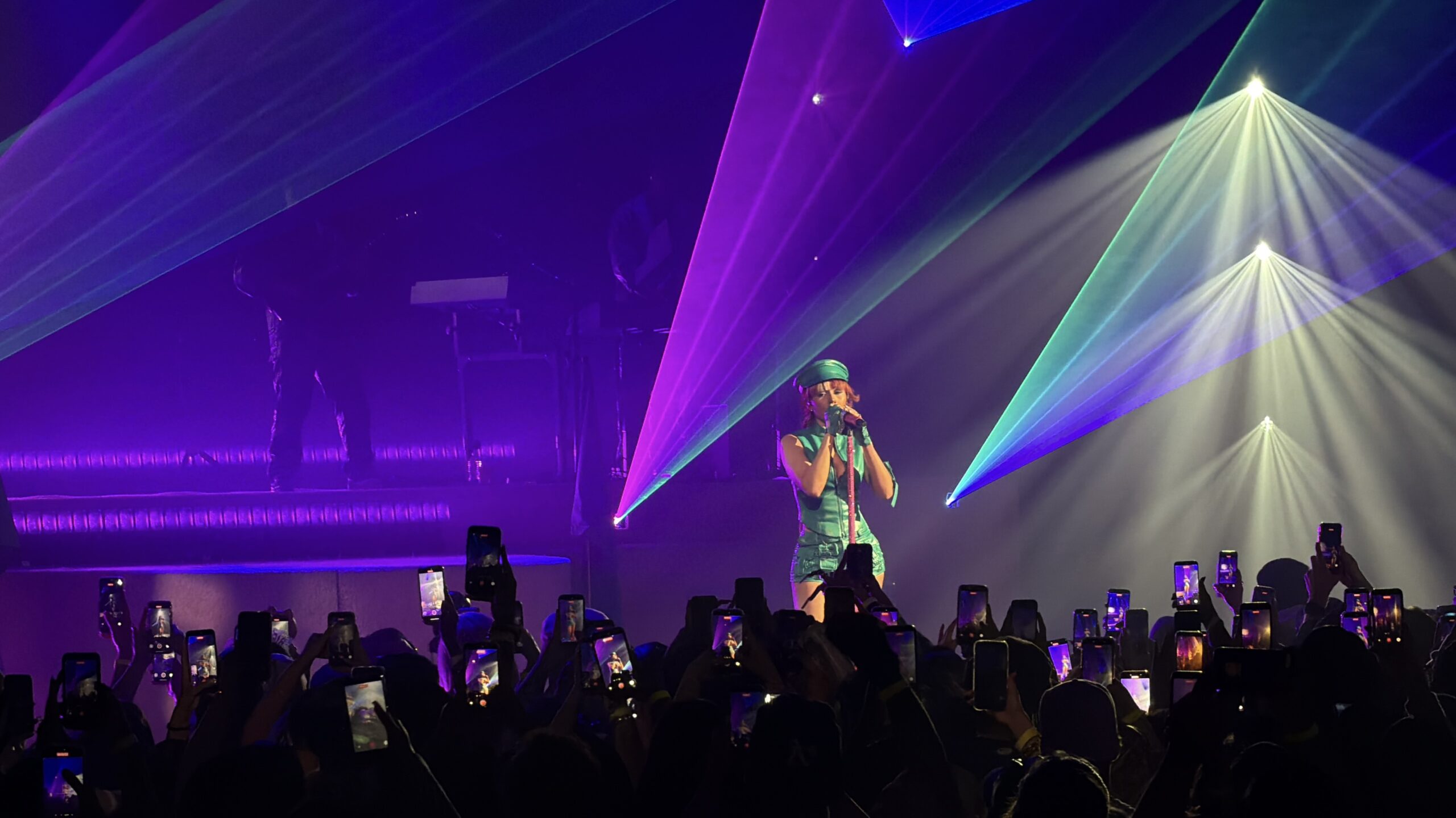 Audience photo of Mariah the Scientist singing beneath purple, green, and blue laser beams as the crowd films on phones.
