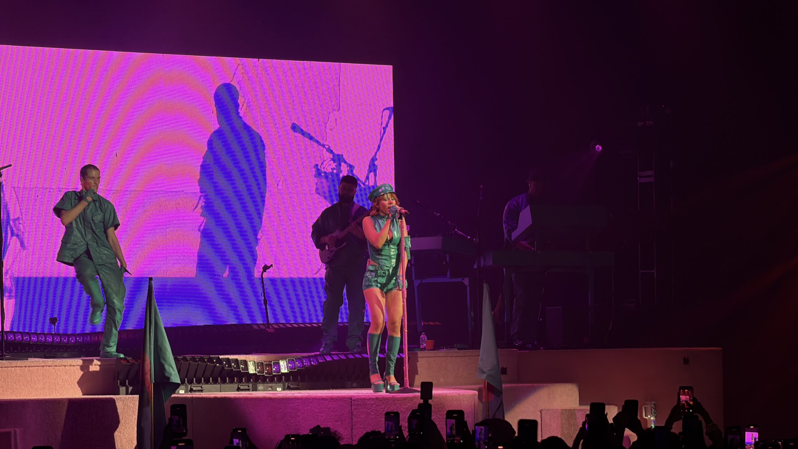 Mariah the Scientist performs in a green toy-soldier inspired outfit on the Hearts Sold Separately Tour at Arizona Financial Theatre in Phoenix, framed by a soft pink stage screen and her band in the background.