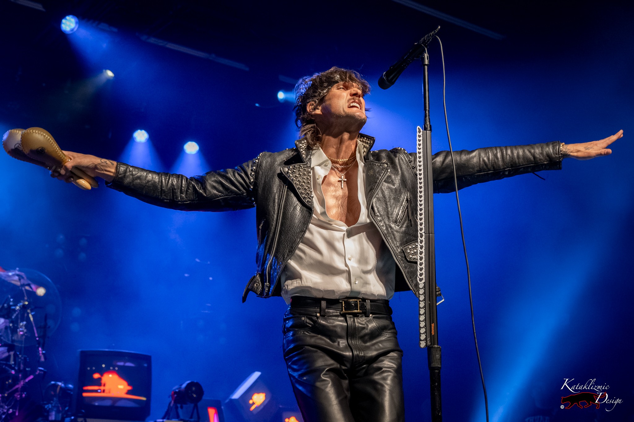 BOYS LIKE GIRLS frontman Martin Johnson stretches his arms wide onstage while holding maracas under blue concert lights.